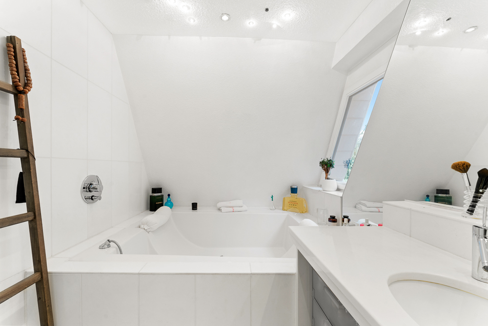 Helles Badezimmer mit Badewanne, großem Spiegel, Fenster und Dekoleiter aus Holz.