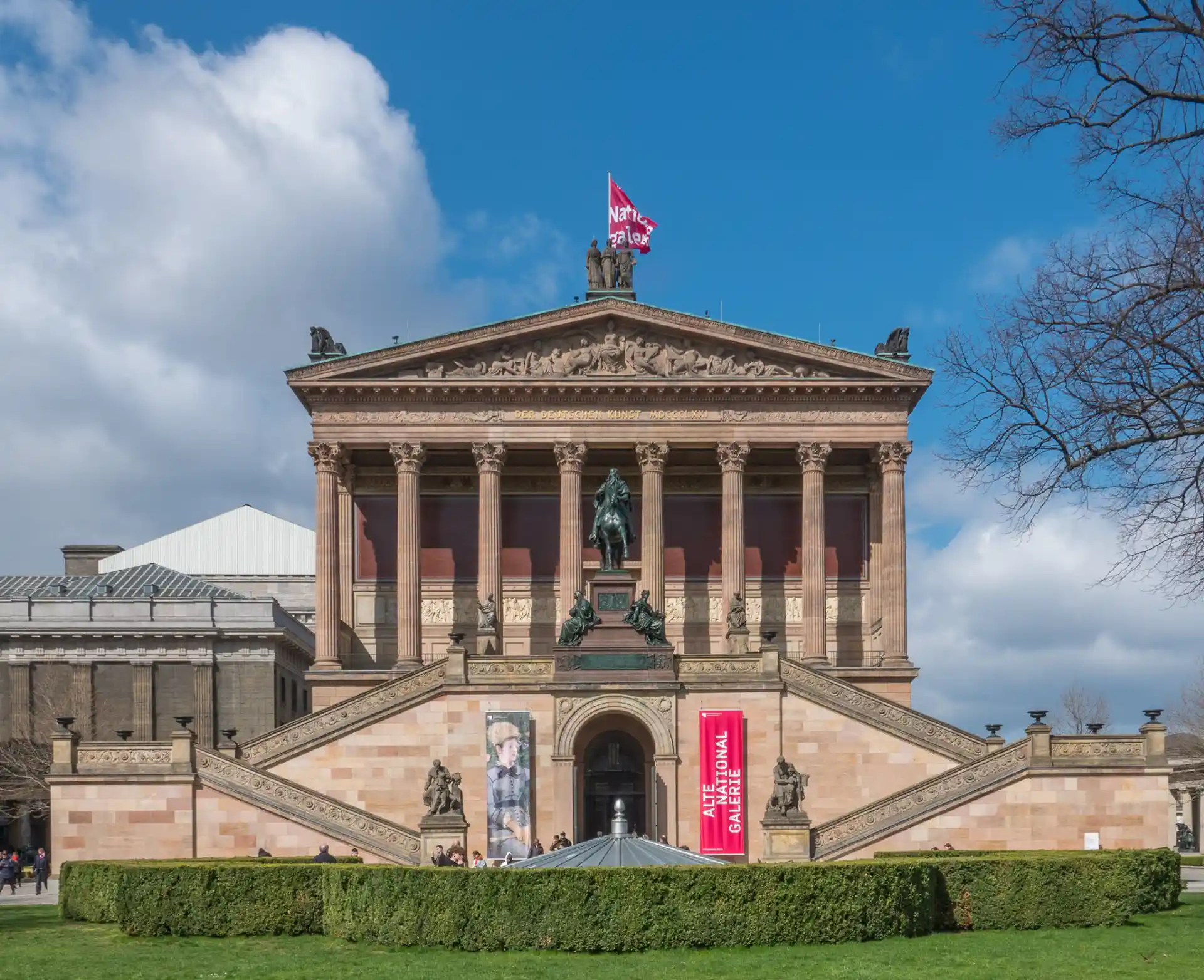 Großes historisches Gebäude mit Säulen, Statuen und Treppen, davor gepflegte Grünanlage.