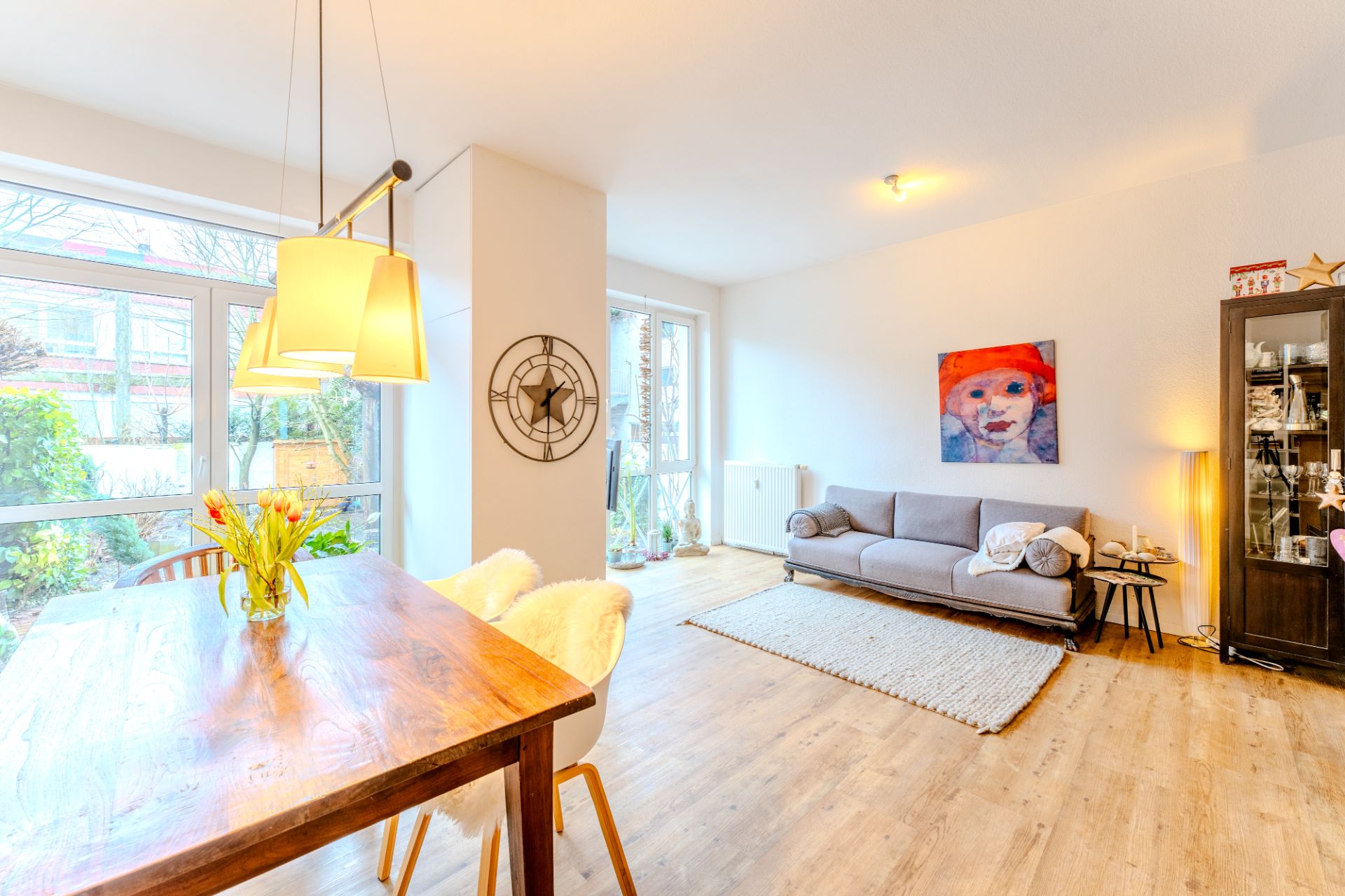 Helles Wohnzimmer mit großem Fenster, Holztisch, grauem Sofa und Wandbild.