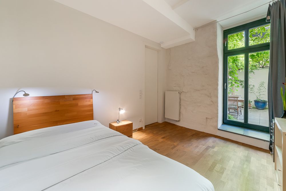 Helles Schlafzimmer mit Holzboden, Bett, Nachttischlampe und Tür zu einem begrünten Innenhof.