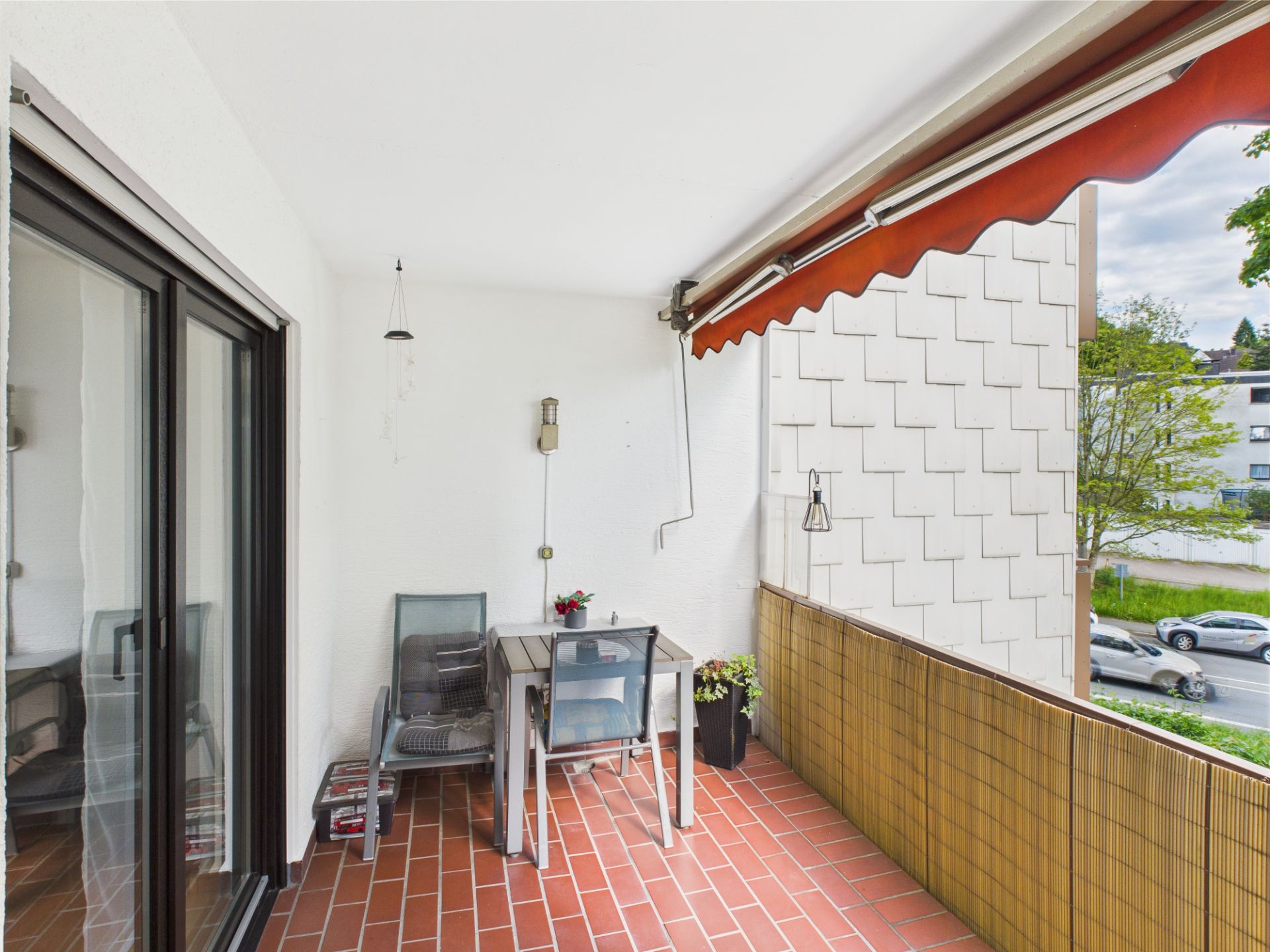 Überdachter Balkon mit zwei Stühlen, Tisch, Blumentöpfen und Markise auf rotem Fliesenboden.