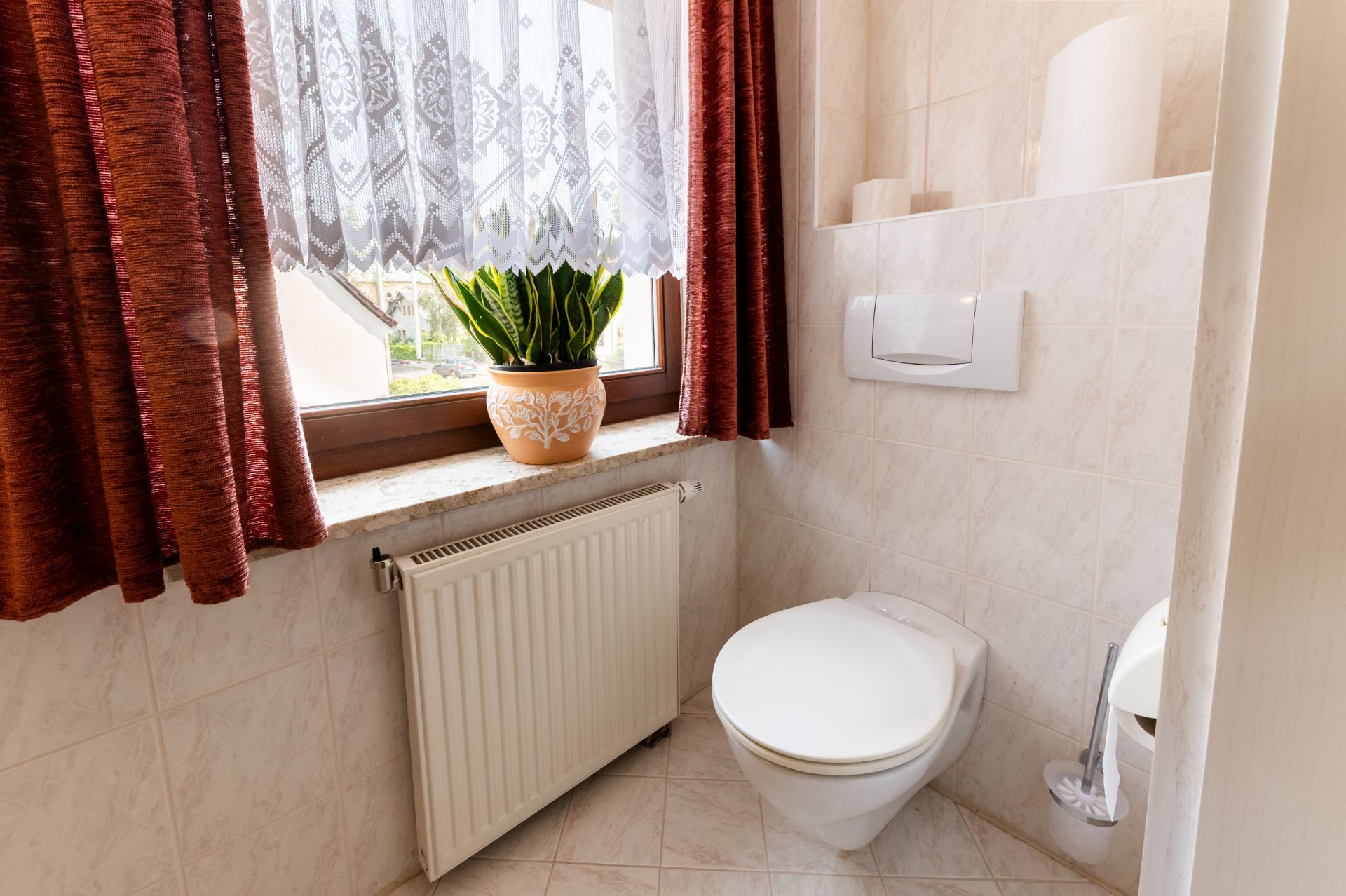 Helles Badezimmer mit Wandtoilette, Fenster, Topfpflanze und roten Vorhängen.