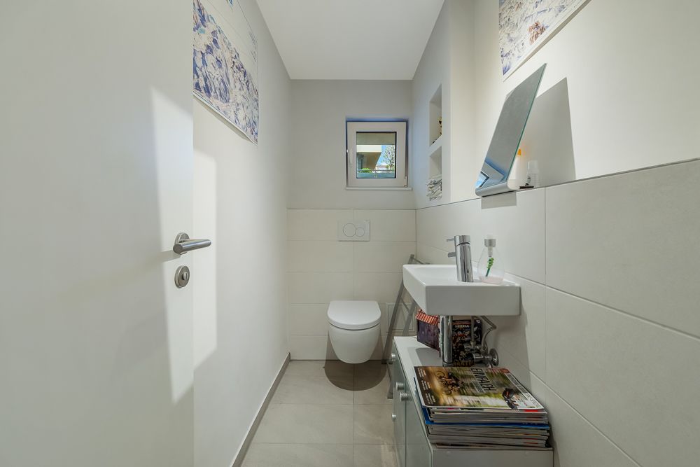 Kleines, modernes Badezimmer mit WC, Waschbecken, Fenster und Zeitschriften auf einem Schrank.