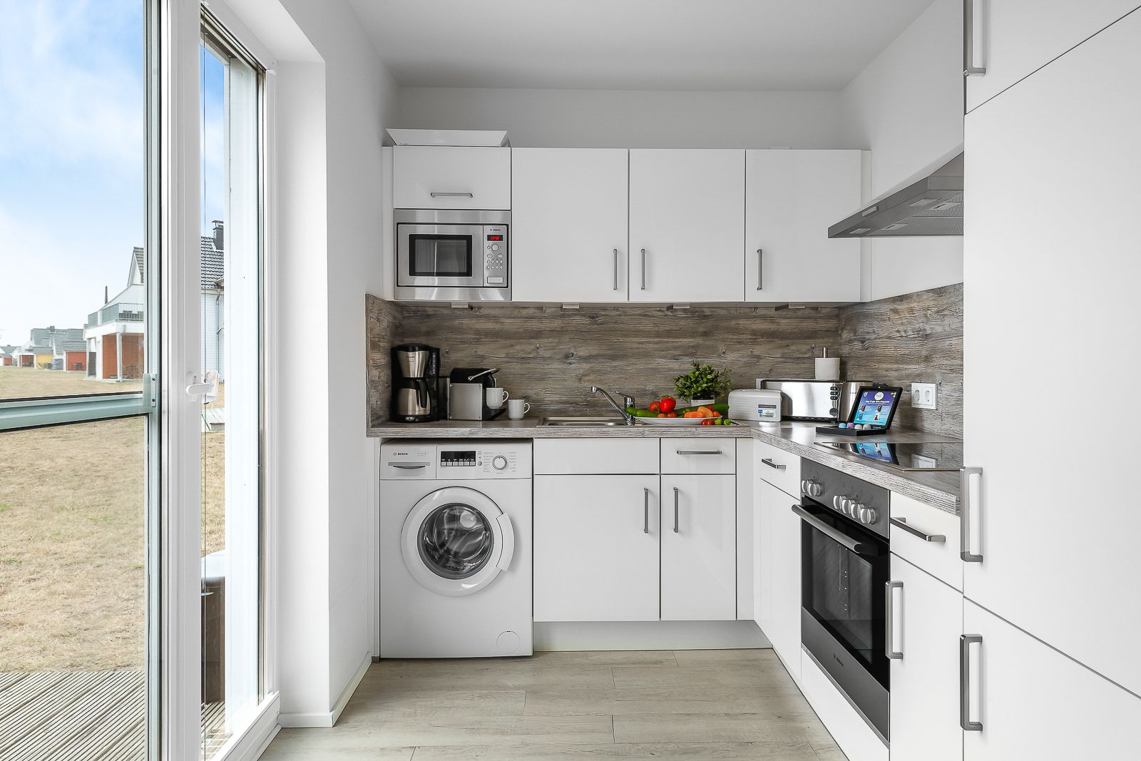 Moderne, helle Küche mit weißen Schränken, Waschmaschine, Ofen und großen Fenstern.