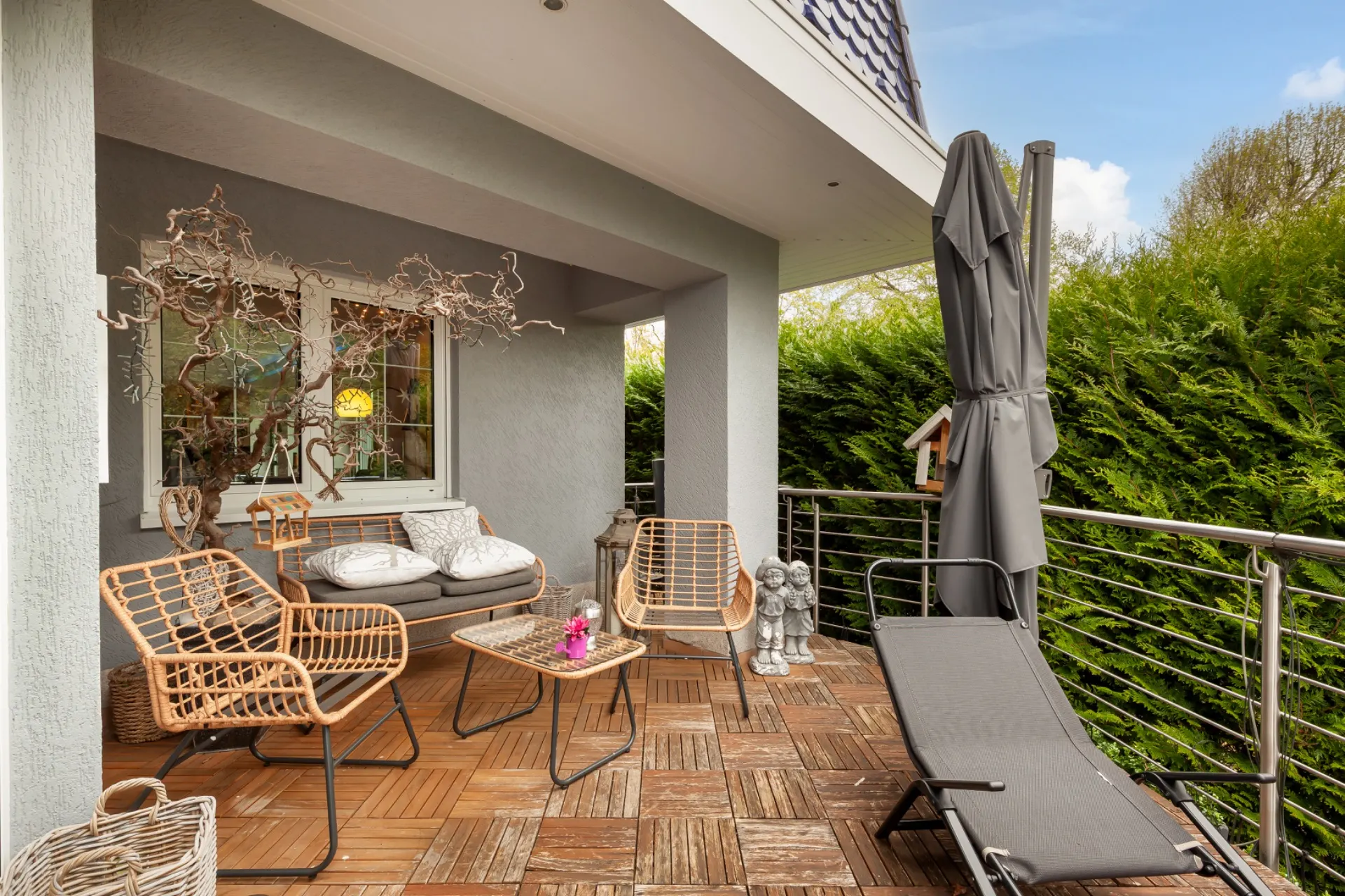 Überdachte Terrasse mit Rattanmöbeln, Liege, Tisch und Blick auf grüne Hecke.