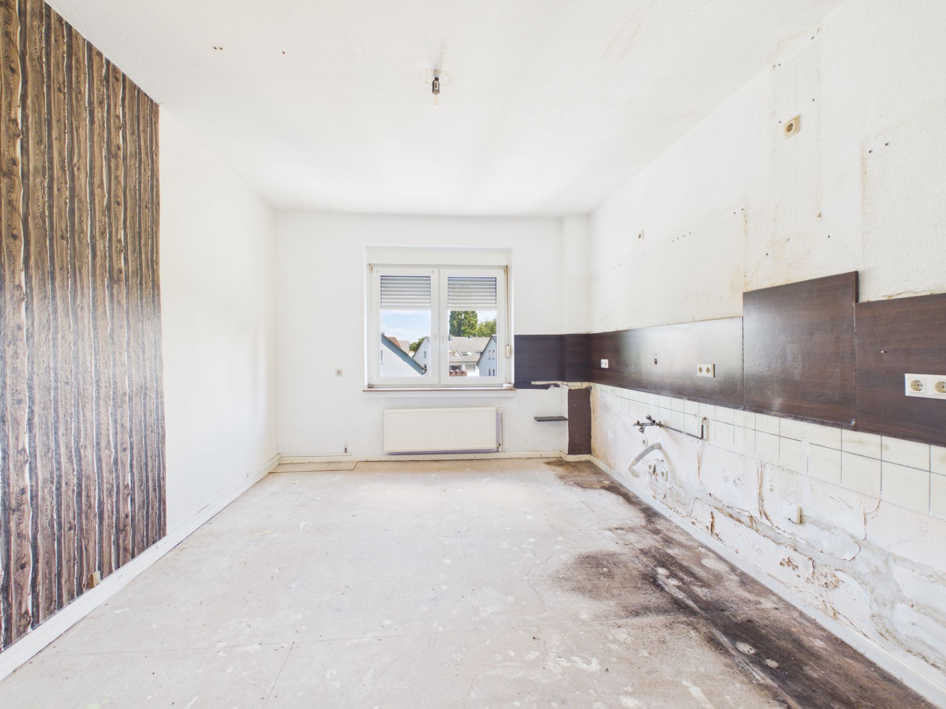 Leerer, renovierungsbedürftiger Raum mit Fenster und sichtbaren Anschlüssen an der Wand.