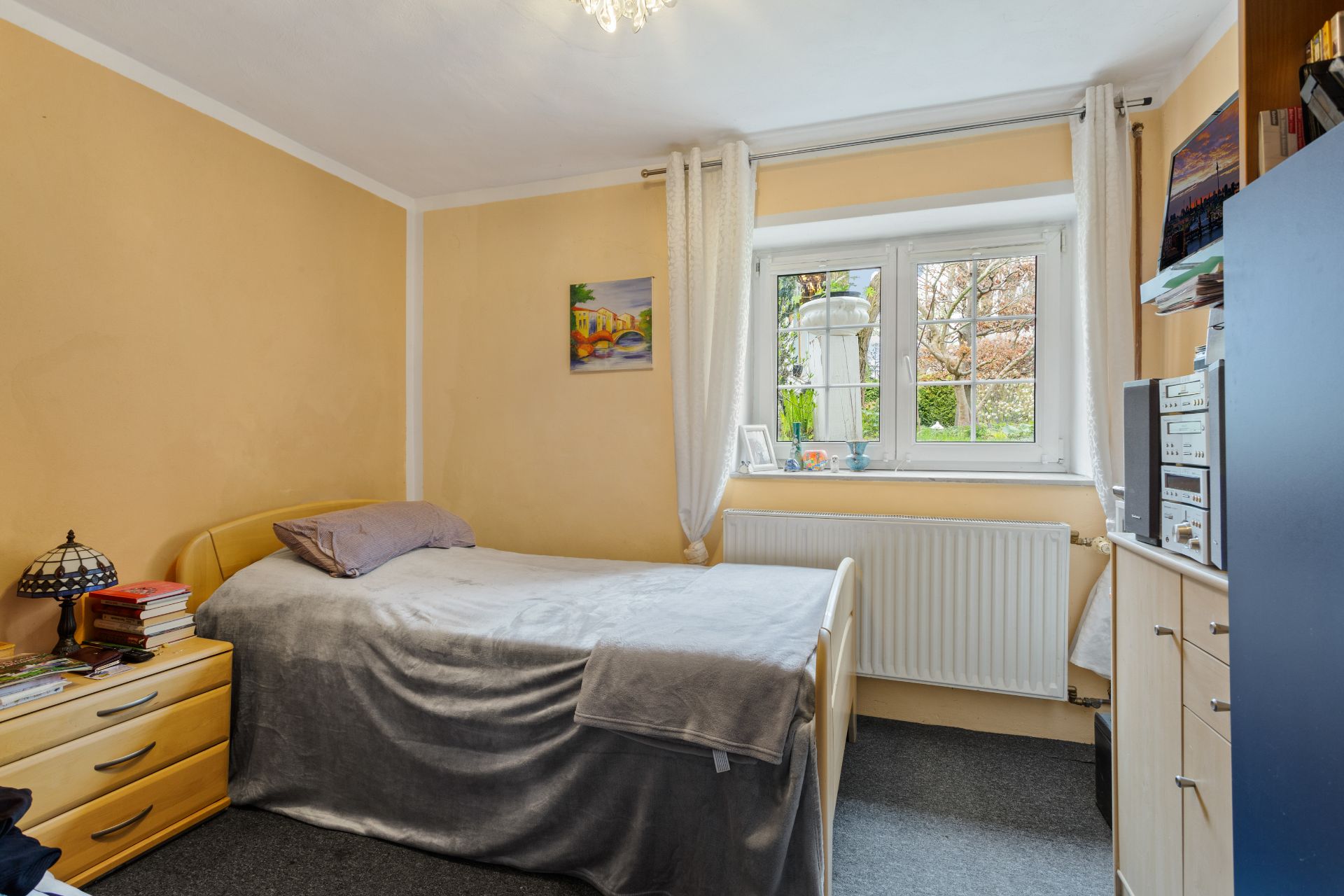 Kleines Schlafzimmer mit Einzelbett, Nachttisch, Kommode und Fenster mit Blick ins Grüne.