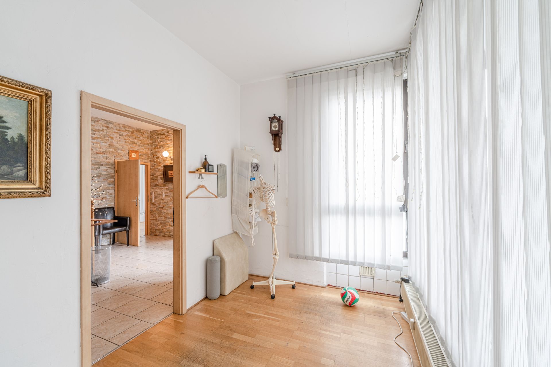 Heller Raum mit Holzboden, Skelettmodell, Ball und großen Fenstern mit Lamellenvorhängen.