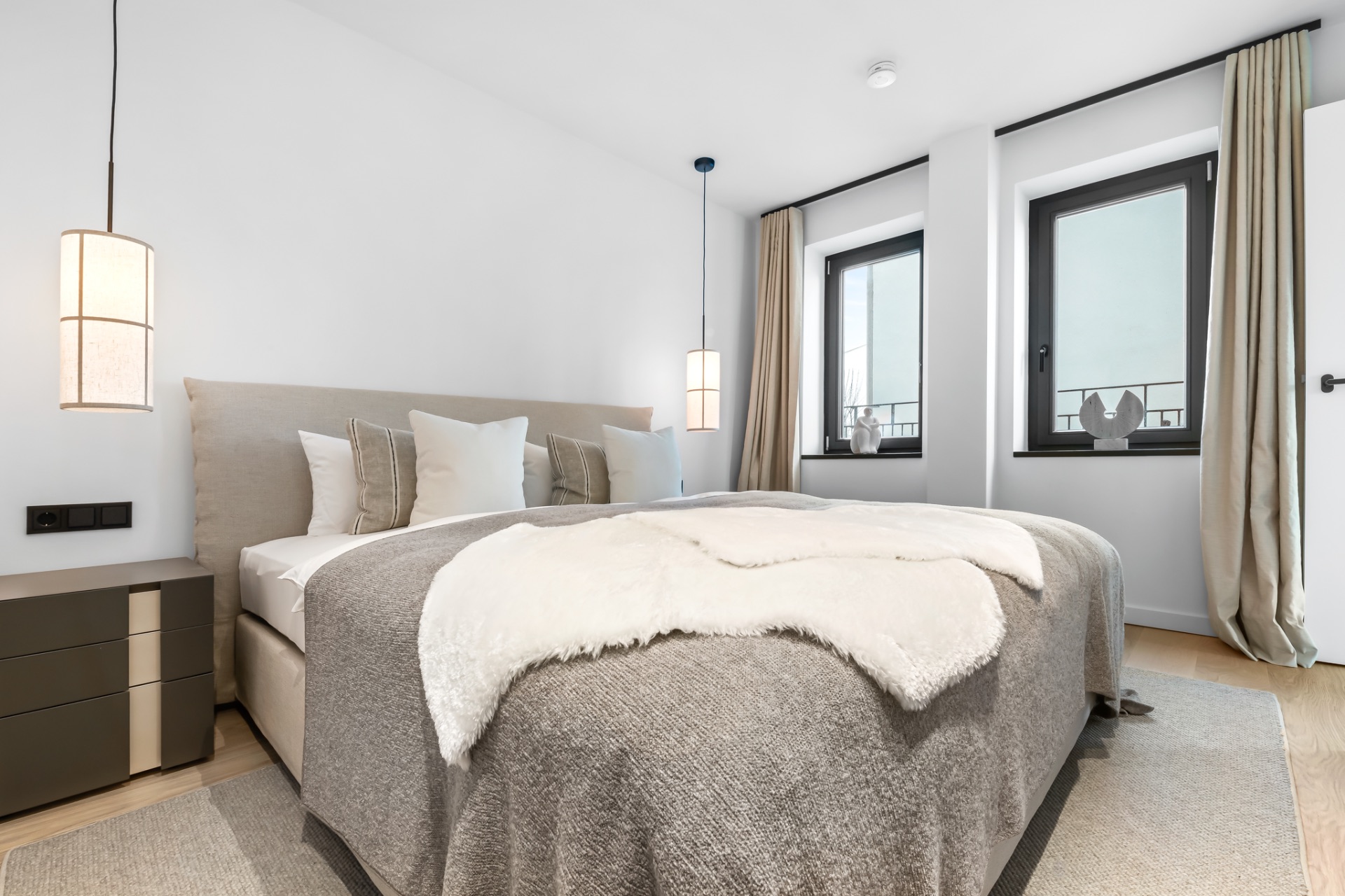 Modernes Schlafzimmer mit Doppelbett, Nachttischen, zwei Fenstern und heller Bettdecke.