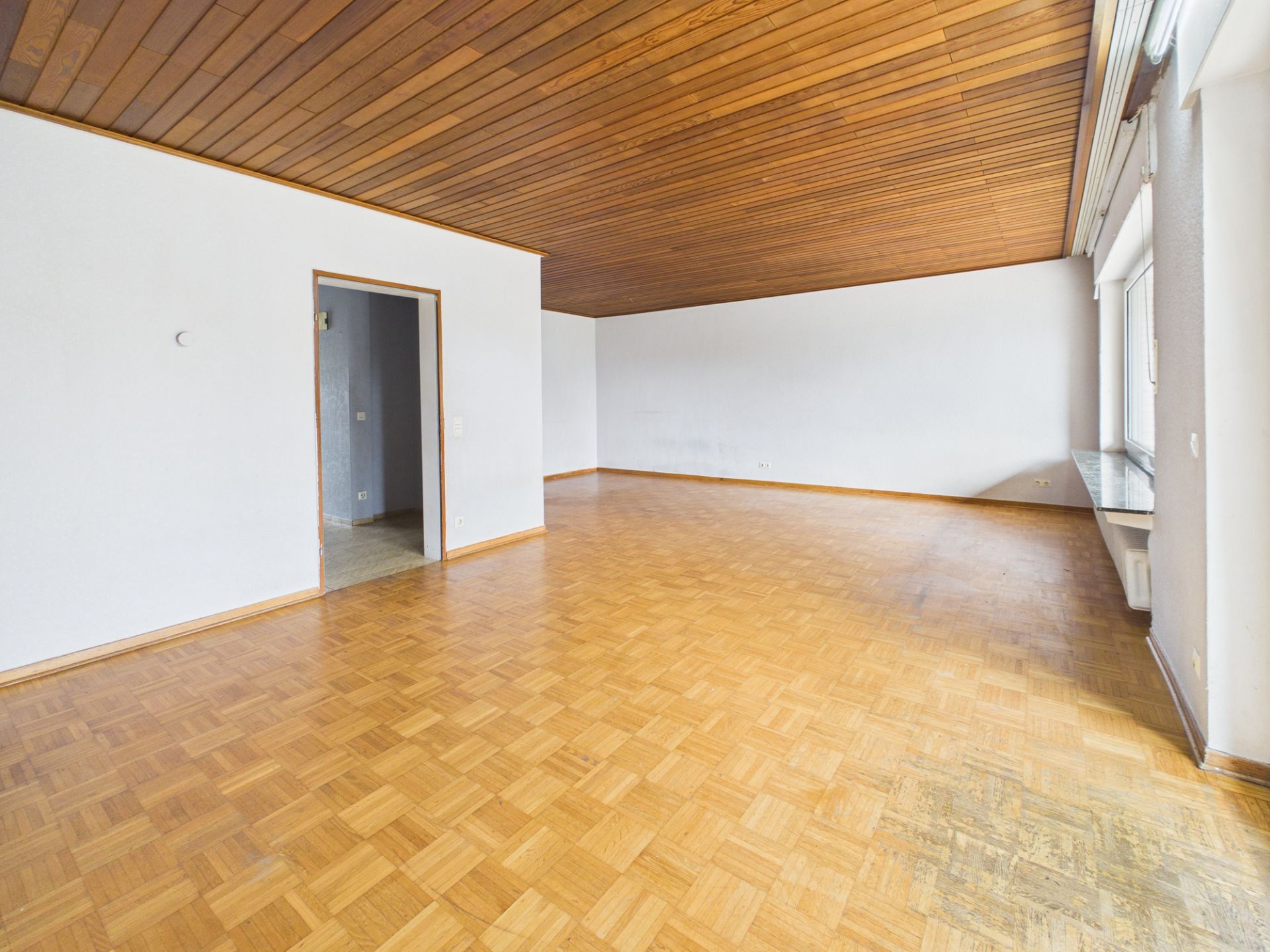 Leerer Raum mit Parkettboden, weißem Wänden, Holzdecke und großem Fenster an der rechten Wand.