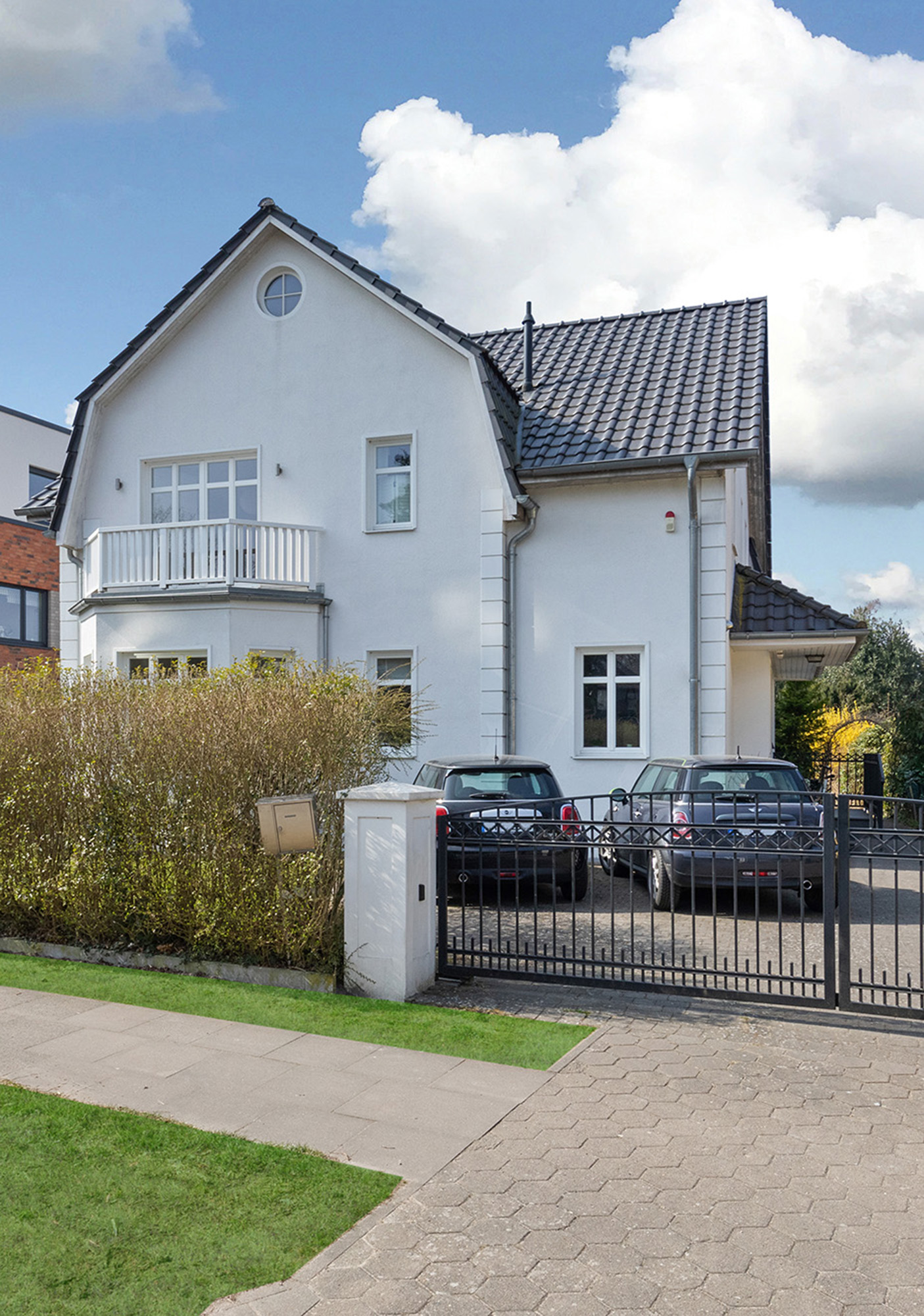 Weißes Einfamilienhaus mit Balkon, Einfahrtstor und zwei geparkten Autos vor dem Haus.