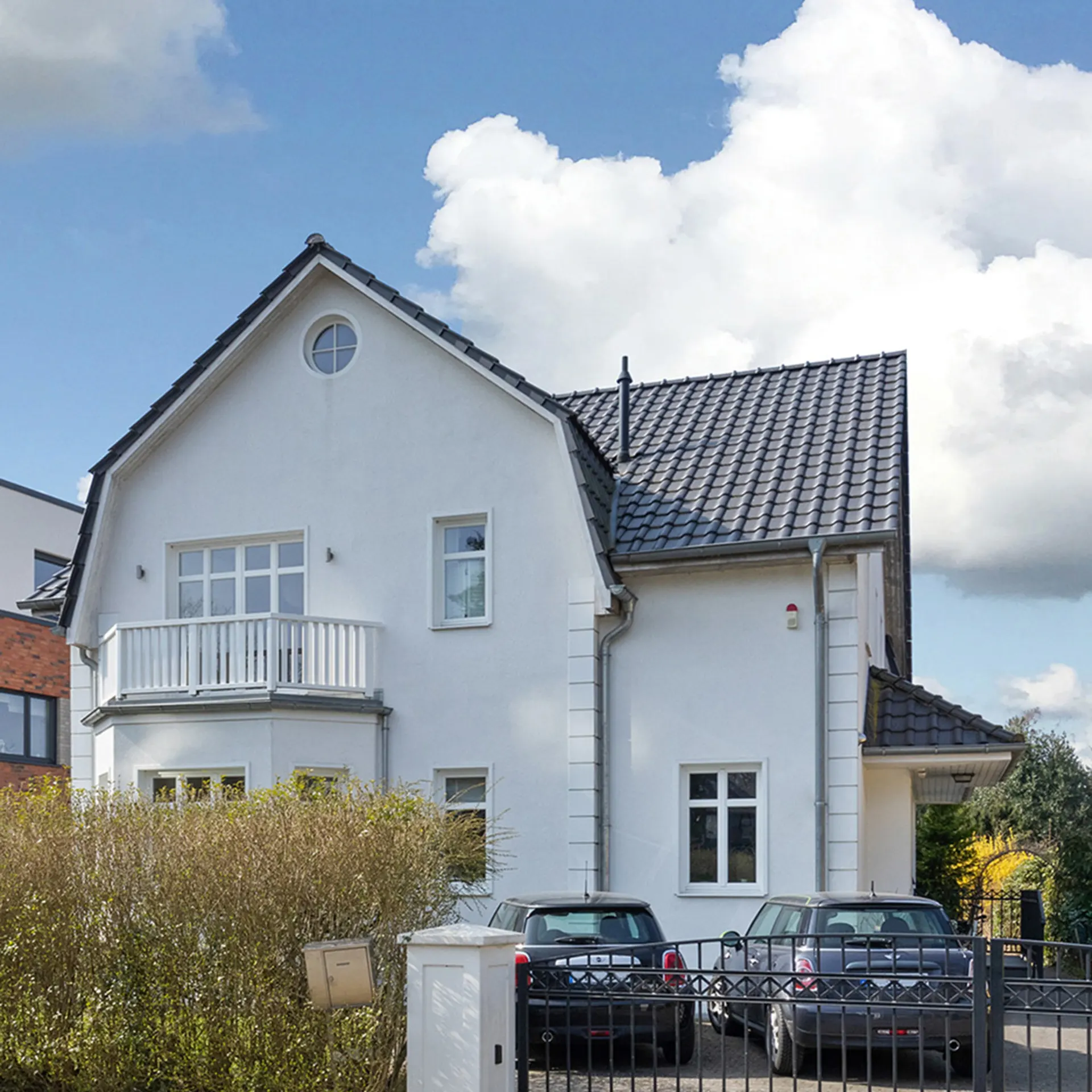 Weißes Einfamilienhaus mit Balkon, Einfahrtstor und zwei geparkten Autos vor dem Haus.