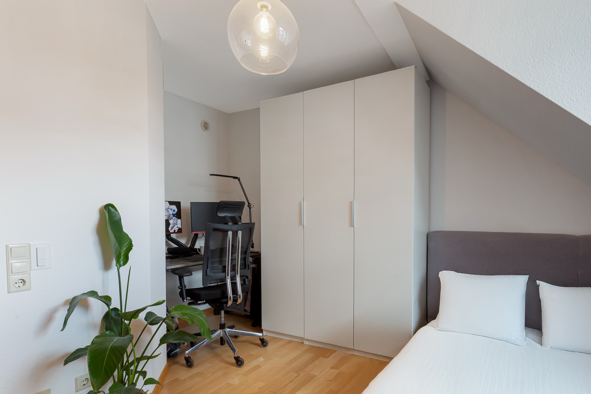 Helles Schlafzimmer mit Bett, weißem Kleiderschrank, Schreibtisch und grüner Pflanze.
