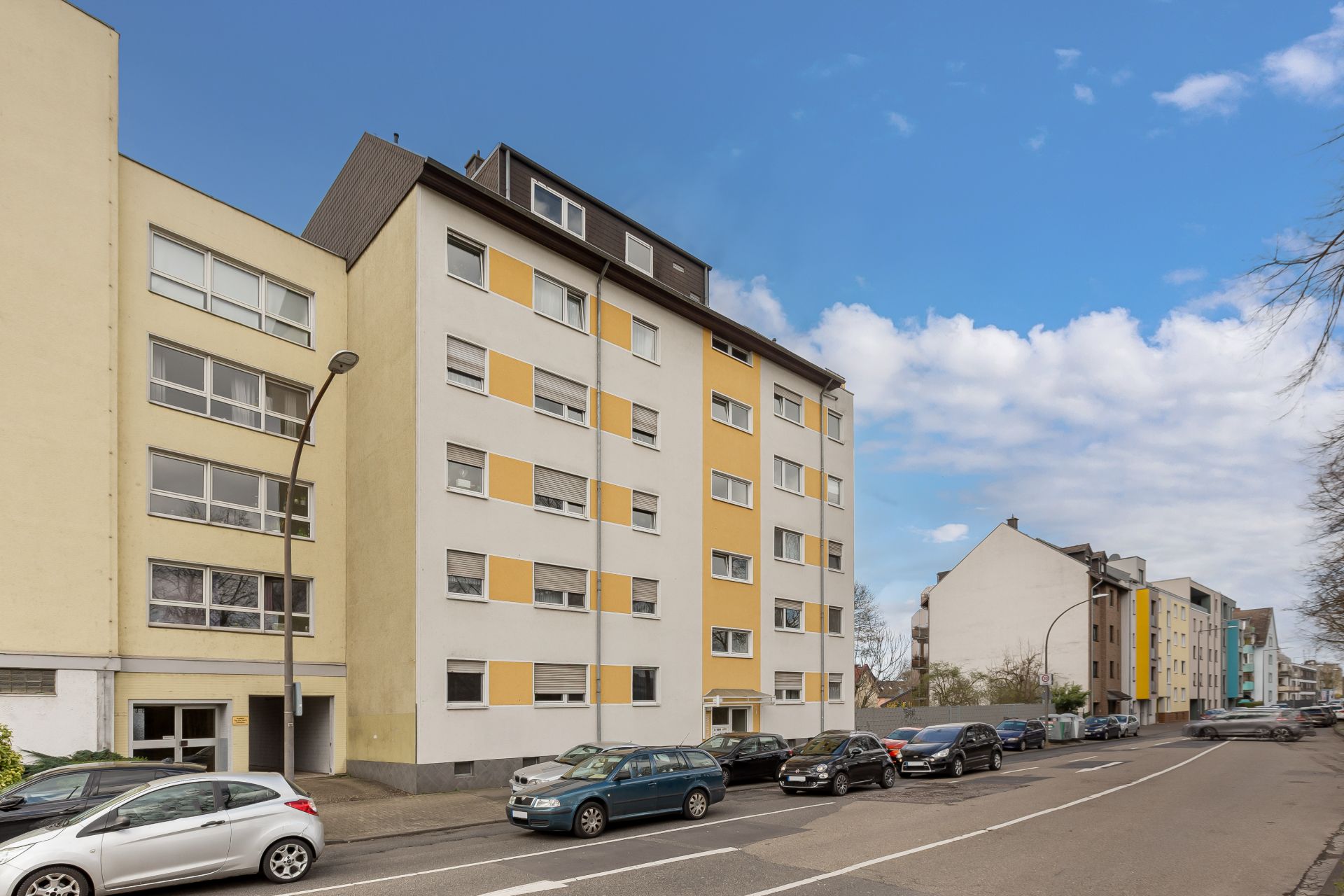 Mehrstöckiges Wohngebäude mit gelben Elementen an einer Straße mit parkenden Autos.