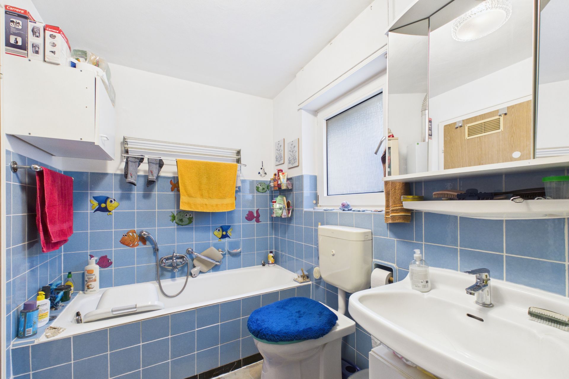 Kleines Badezimmer mit Badewanne, Waschbecken, Toilette und blauen Fliesen mit bunten Fischen.