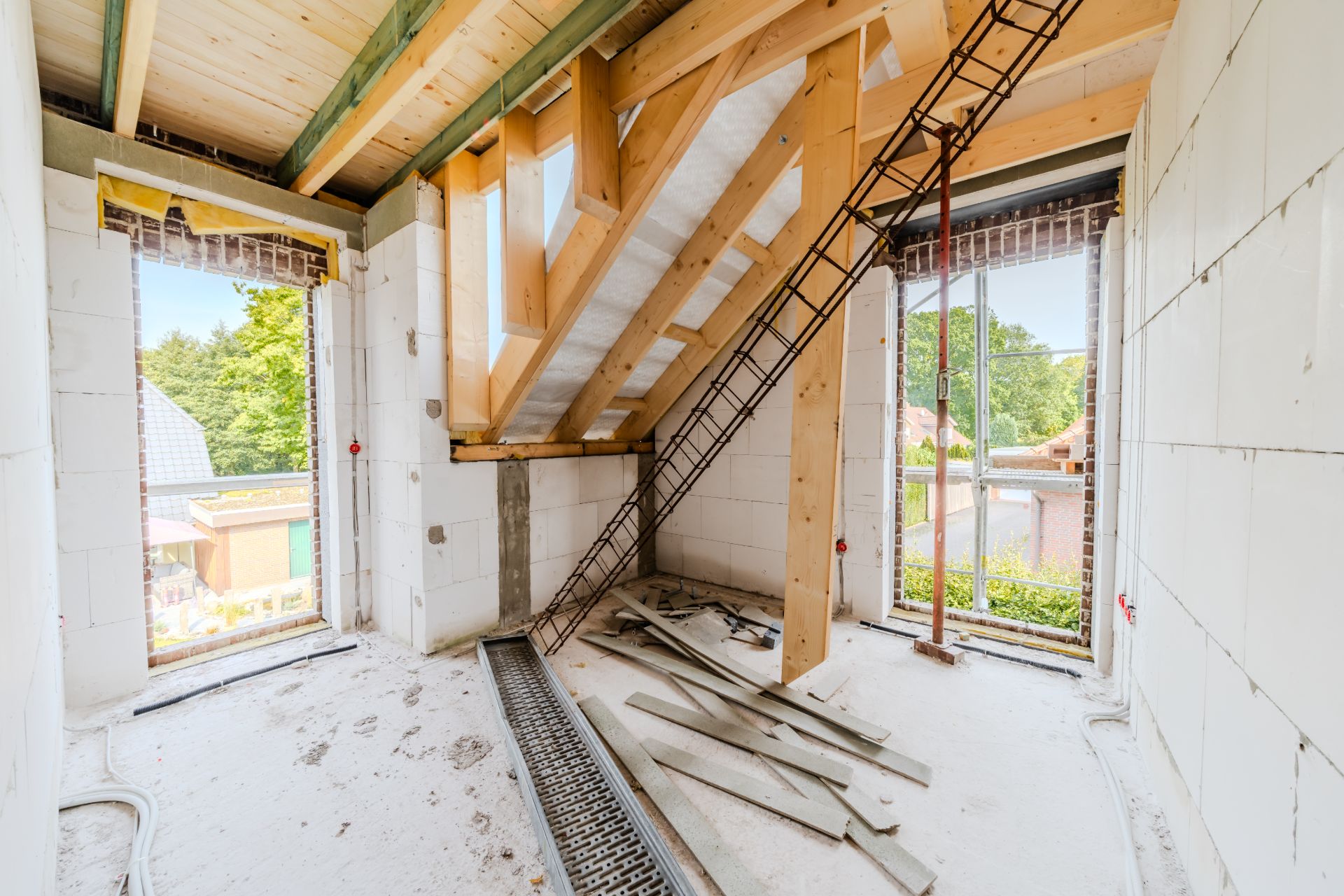 Ein Raum im Rohbau mit Holzbalken, offenen Fenstern und Baumaterial auf dem Boden.