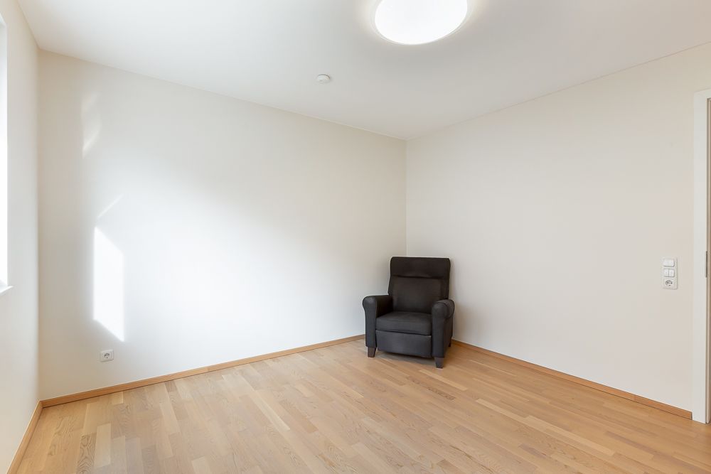 Helles, leeres Zimmer mit Holzboden und einem schwarzen Sessel in der Ecke.