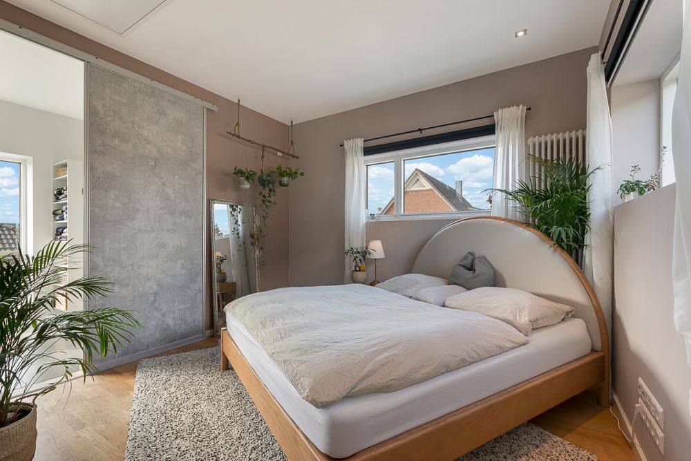 Modernes Schlafzimmer mit großem Bett, Pflanzen und Fenstern mit weißen Vorhängen.