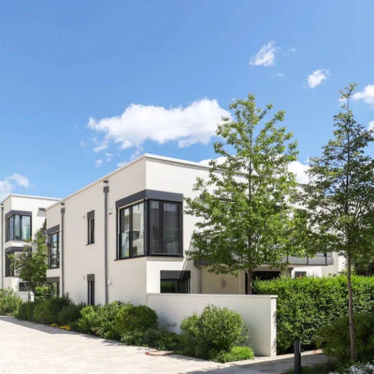 Moderne Wohnhäuser mit großen Fenstern, Bäumen und gepflegten Grünflächen bei Sonnenschein.