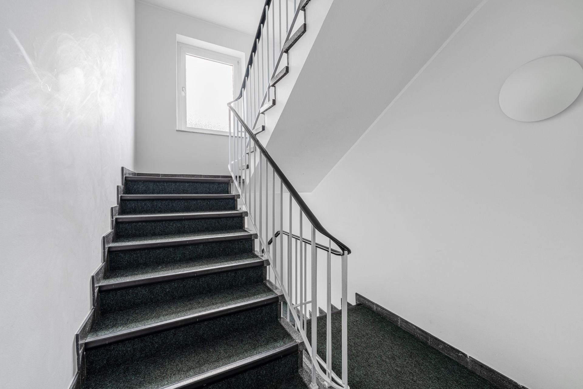 Innen-Treppenhaus mit dunklem Teppich und weißem Geländer, Tageslicht durch Fenster.