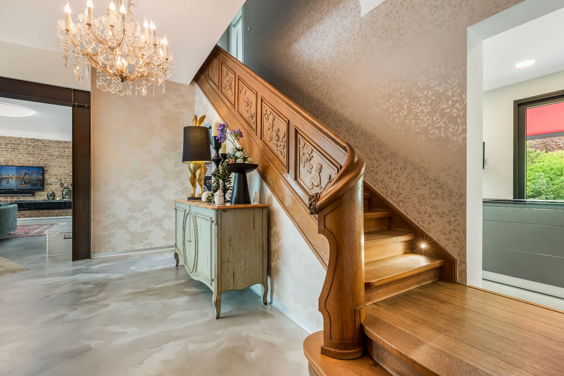 Holztreppe mit geschnitztem Geländer, Kronleuchter und Kommode mit Dekoration im Flur.