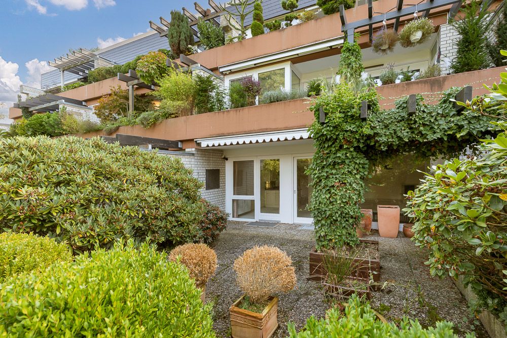 Terrasse mit Pflanzen und Sträuchern vor einer modernen Wohnanlage mit großen Fenstern.