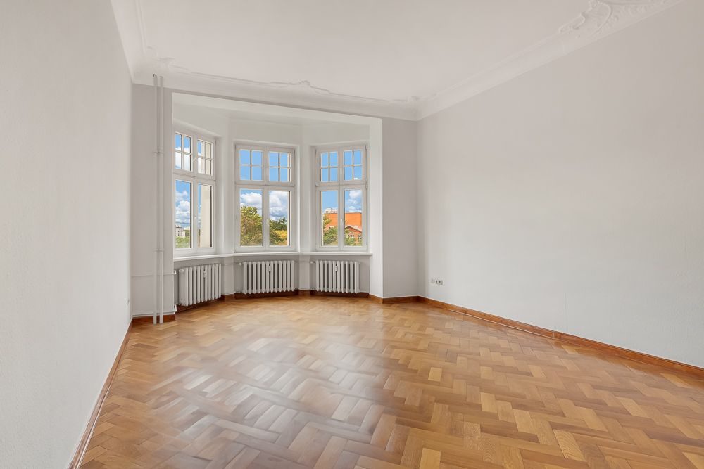 Heller leerer Raum mit Parkettboden und großen Fenstern mit Blick nach draußen.