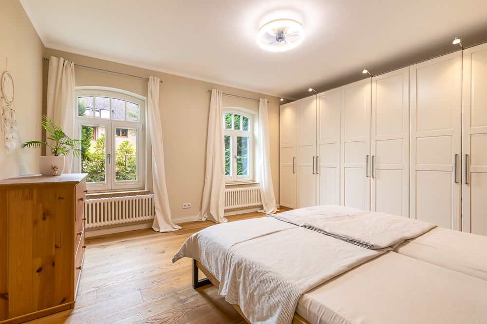 Helles Schlafzimmer mit Doppelbett, weißen Schränken, Kommode und zwei Fenstern mit Vorhängen.