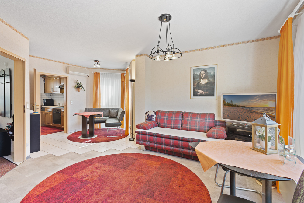 Wohnzimmer mit rotem Sofa, Fernseher, rundem Teppich und Zugang zur Küche.