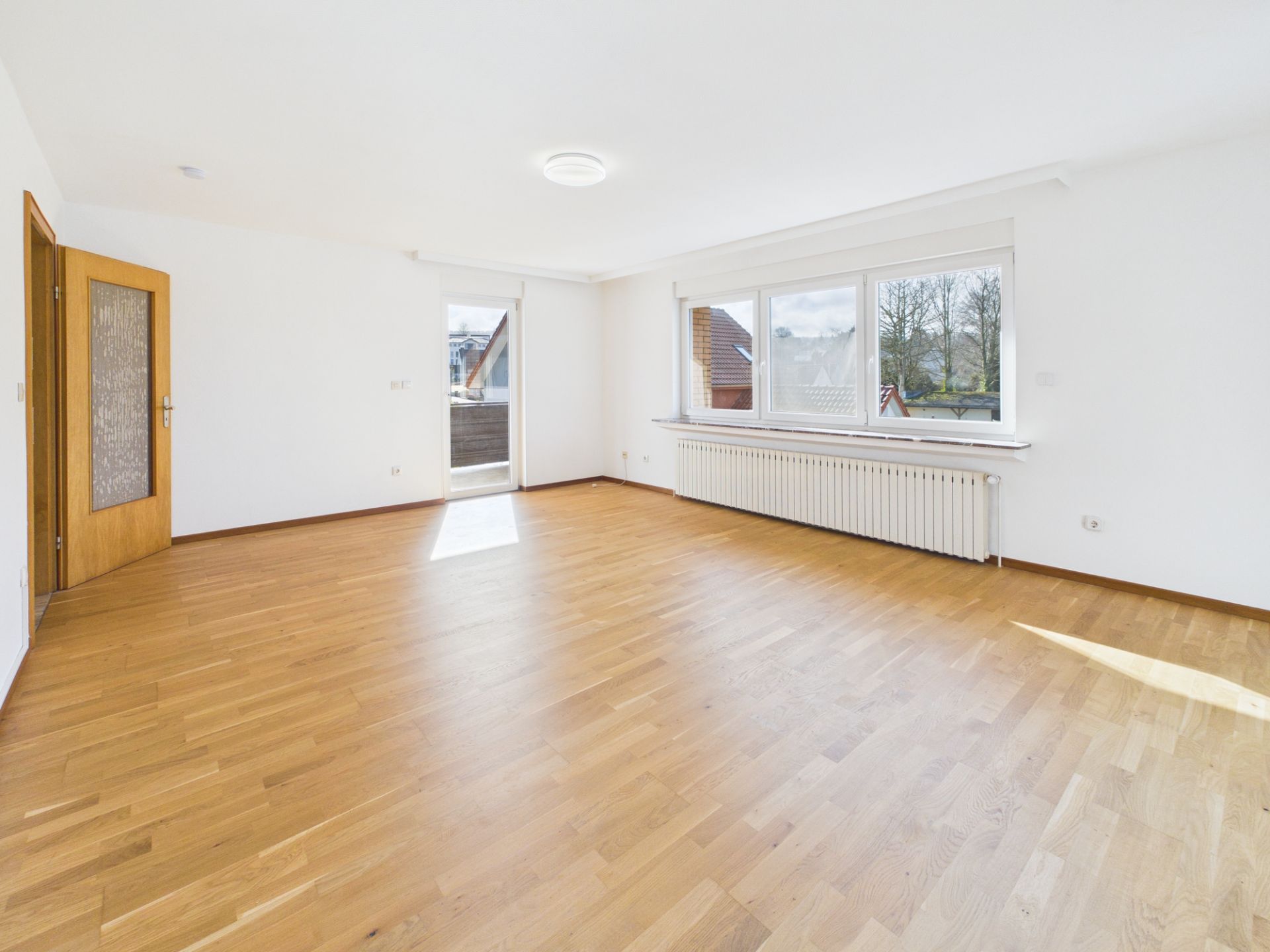 Helles, leeres Zimmer mit Holzfußboden, großer Fensterfront und Tür zum Balkon.