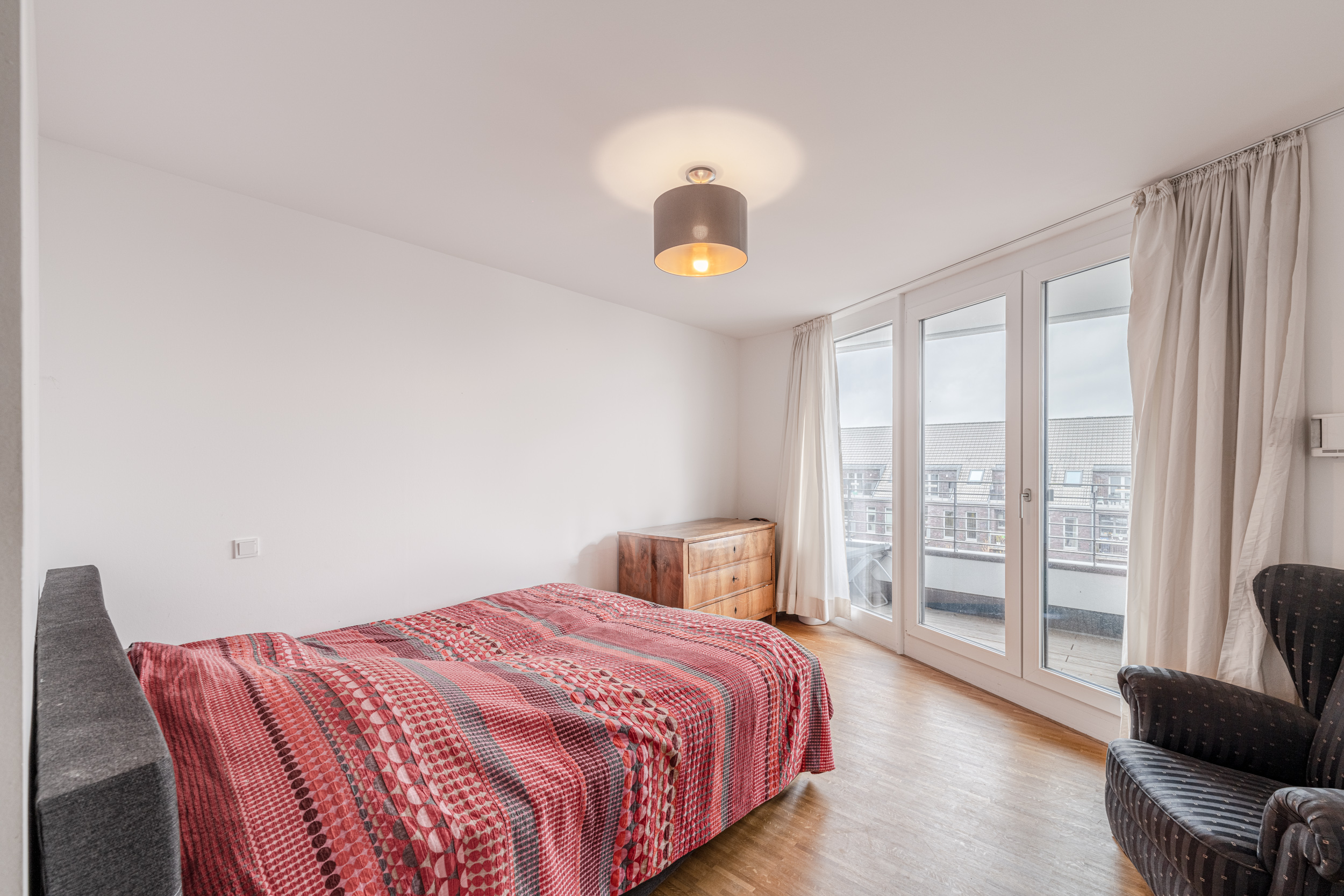 Schlafzimmer mit Doppelbett, bunter Decke, Sessel, Kommode und großen Fenstern mit Vorhängen.