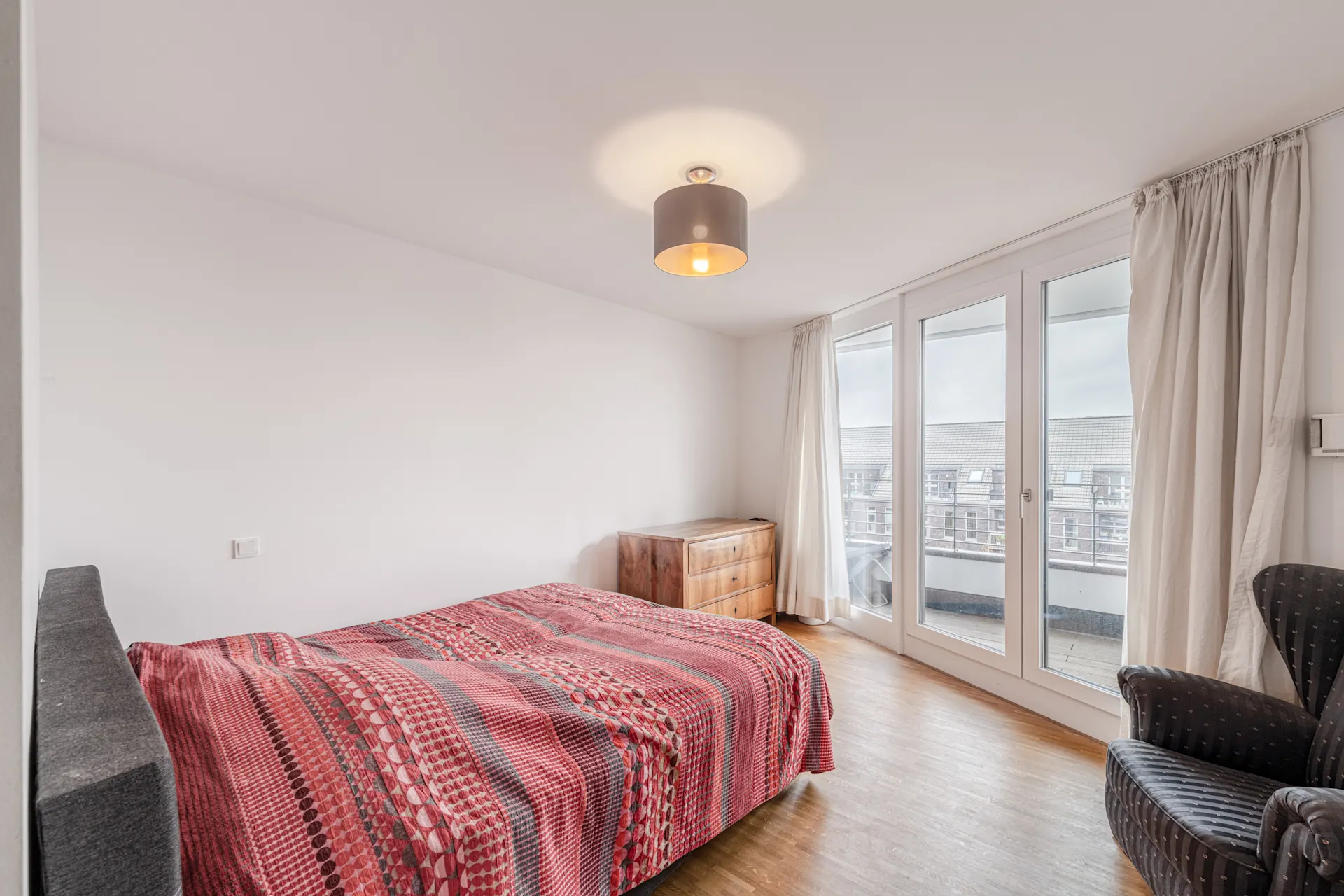 Schlafzimmer mit Doppelbett, bunter Decke, Sessel, Kommode und großen Fenstern mit Vorhängen.