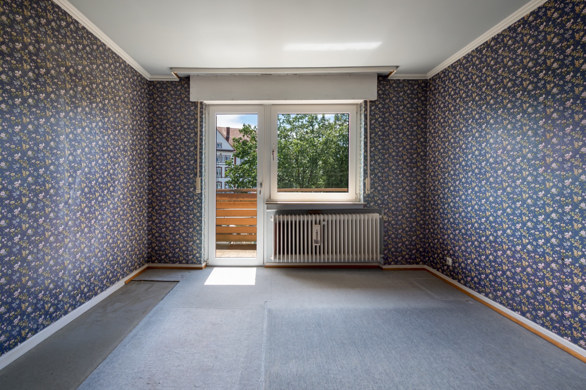 Leeres Zimmer mit gemusterter Tapete, Teppichboden und Tür zu einem kleinen Balkon.