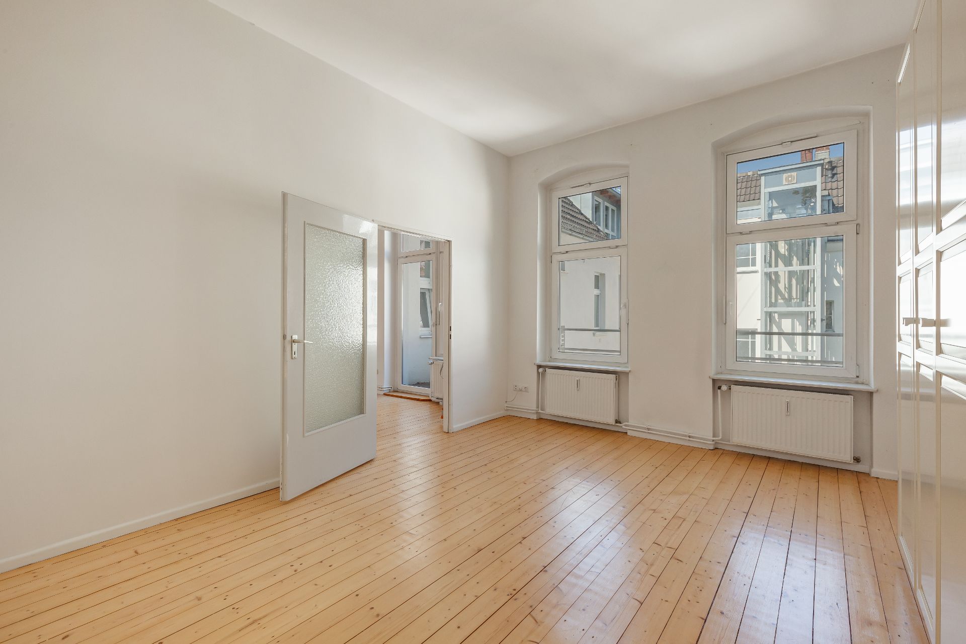Helles, leeres Zimmer mit Holzfußboden, zwei Fenstern und offener Tür.
