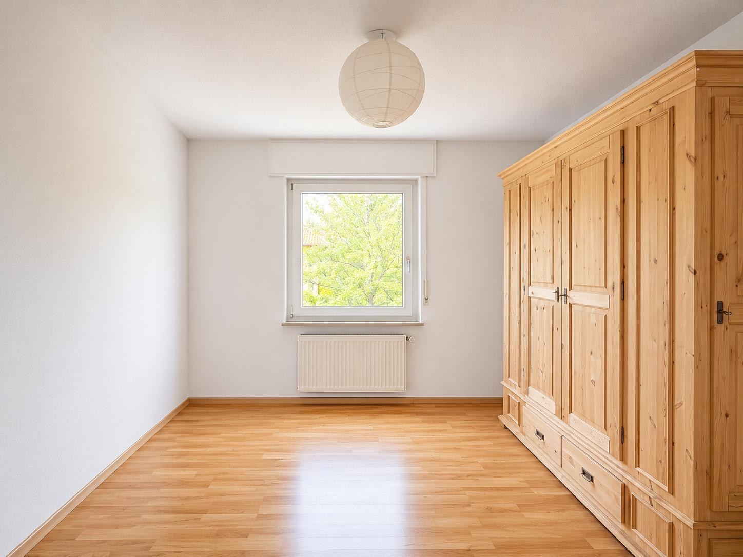 Helles Zimmer mit Holzschrank, Fenster, Heizkörper und Holzfußboden.