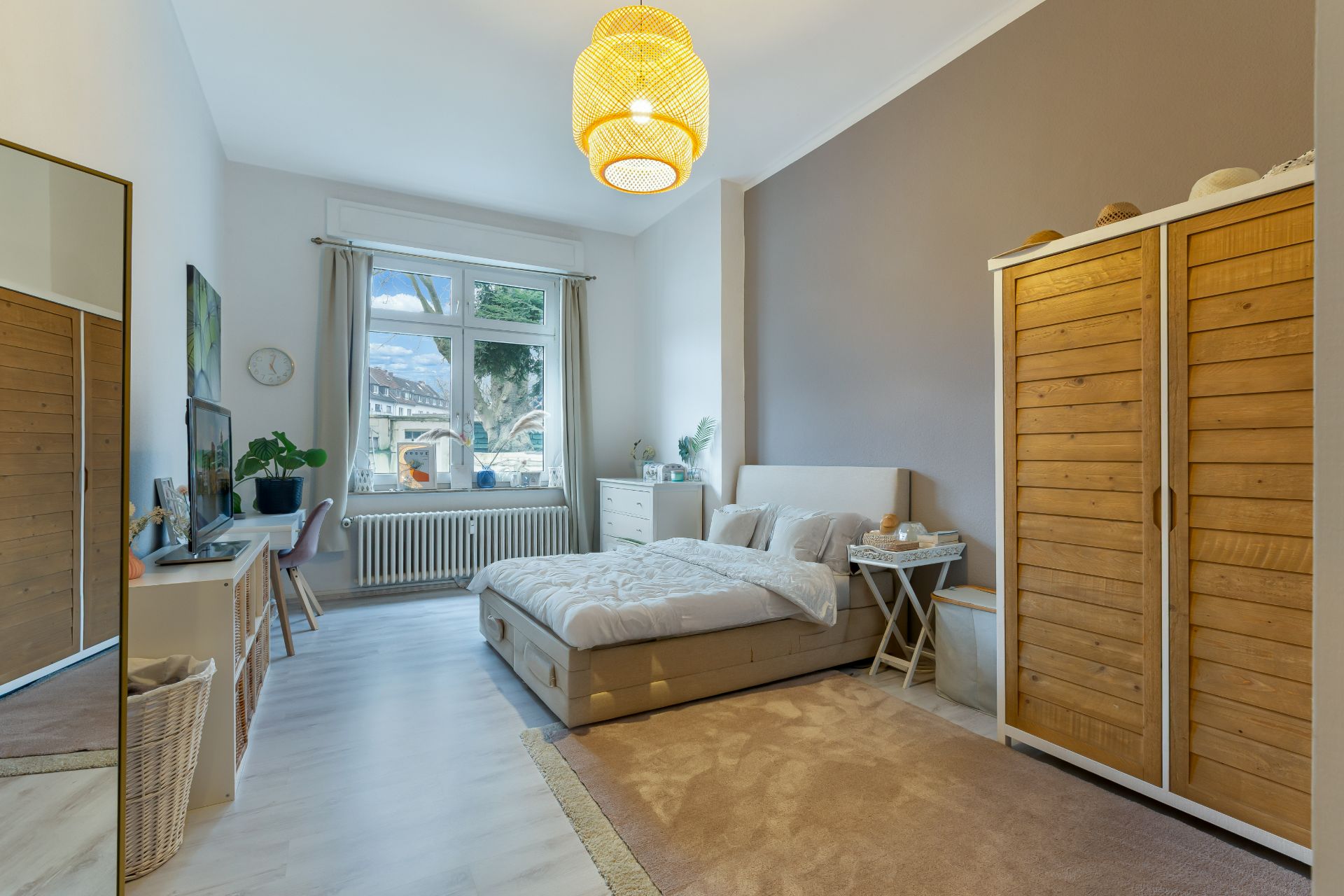 Helles Schlafzimmer mit Doppelbett, Holzschrank, Schreibtisch und großem Fenster.