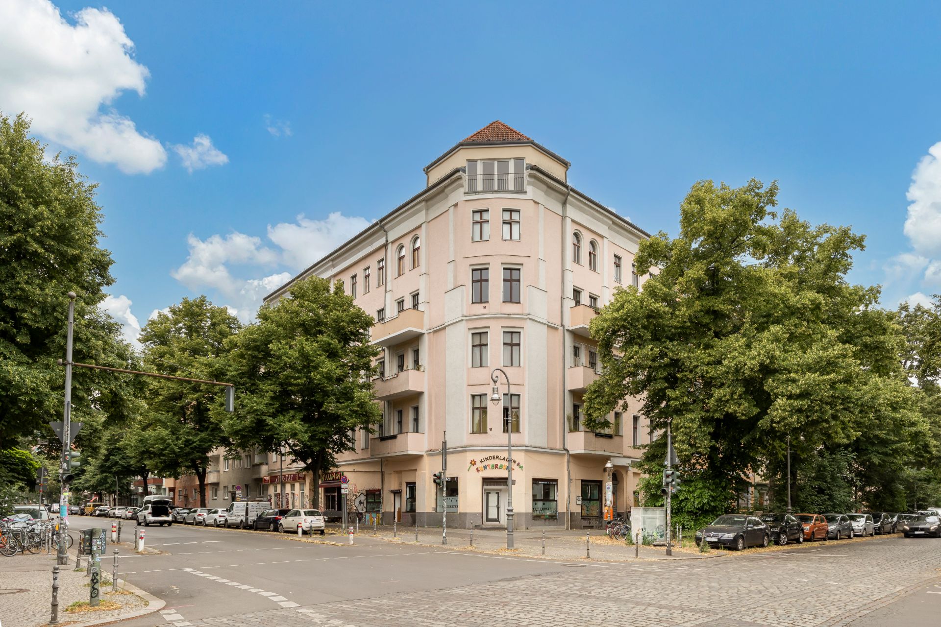 Eckwohnhaus mit mehreren Etagen, Balkonen und Bäumen an beiden Straßenrändern.