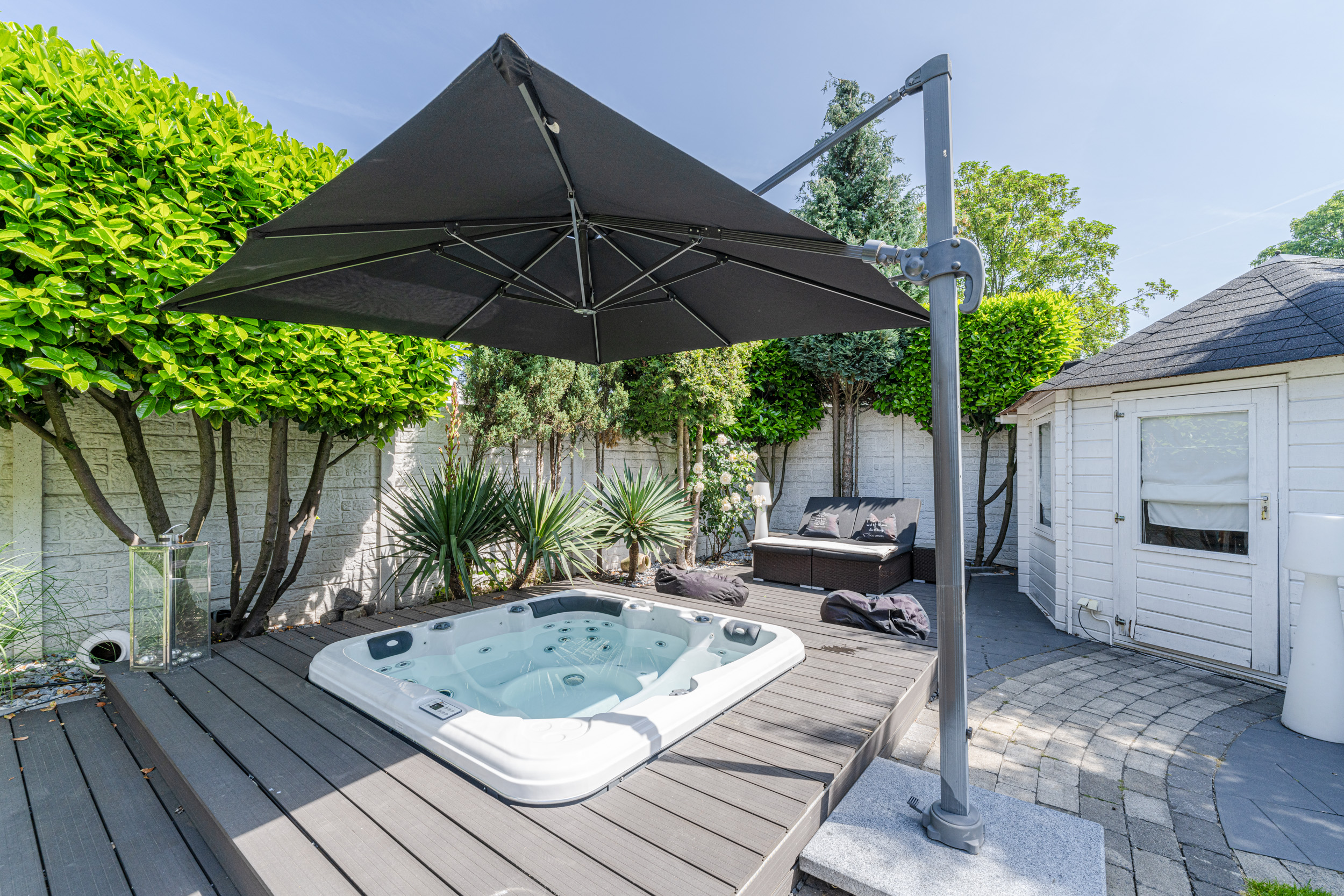 Whirlpool auf Holzterrasse mit großem Sonnenschirm, Gartenmöbeln und weißen Gartenhaus.