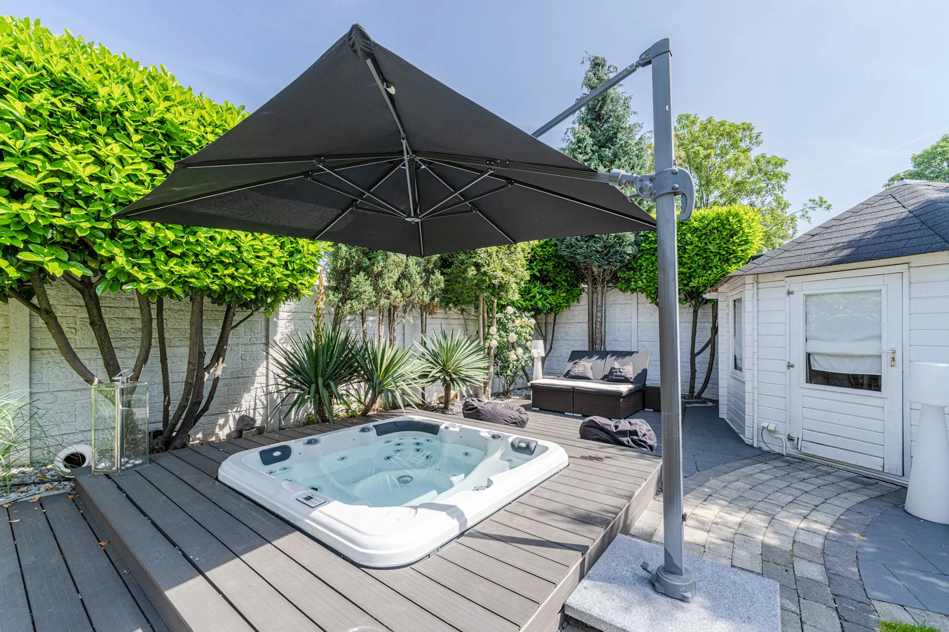 Whirlpool auf Holzterrasse mit großem Sonnenschirm, Gartenmöbeln und weißen Gartenhaus.