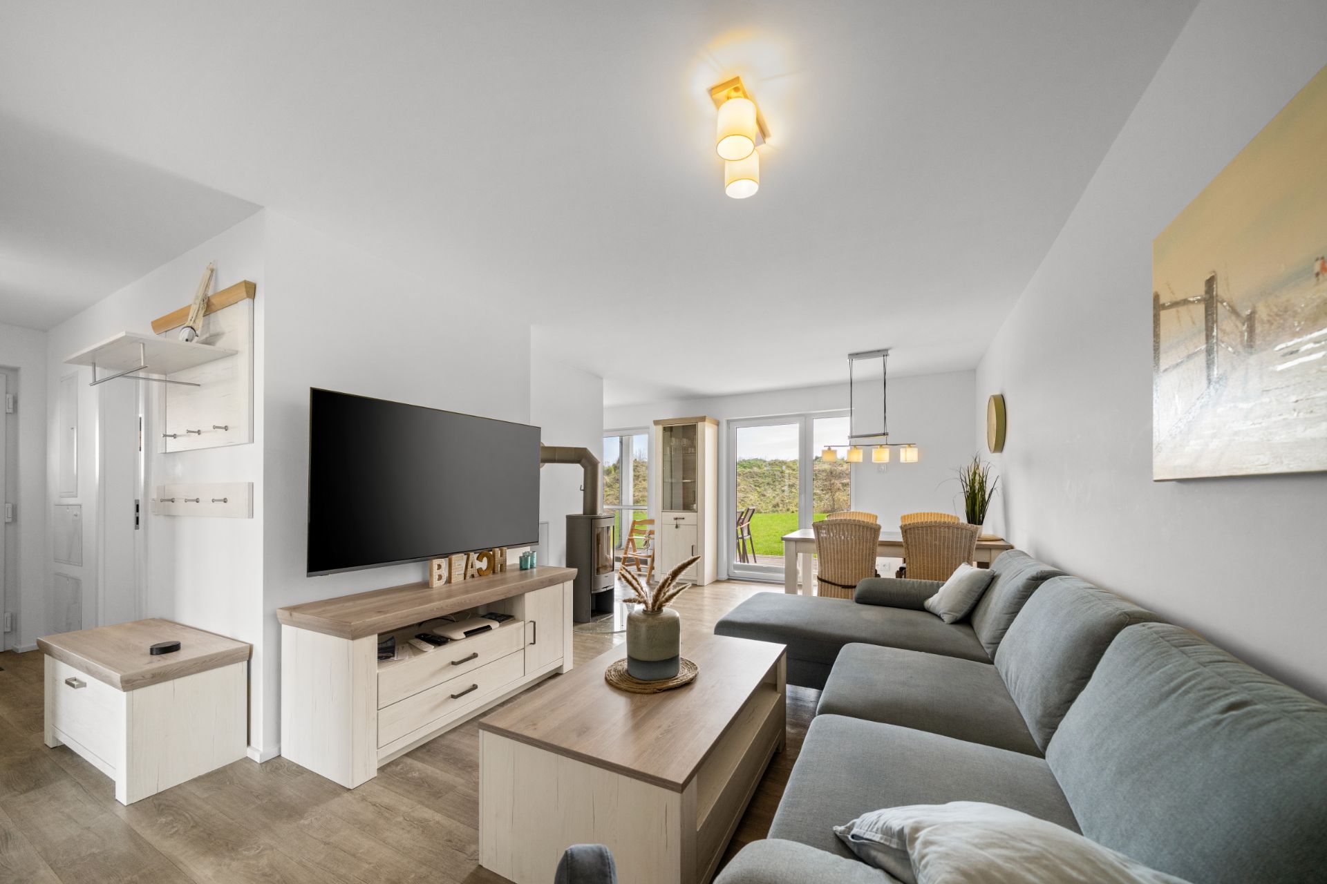 Modernes Wohnzimmer mit grauem Sofa, großem Fernseher und Esstisch, große Fenster im Hintergrund.