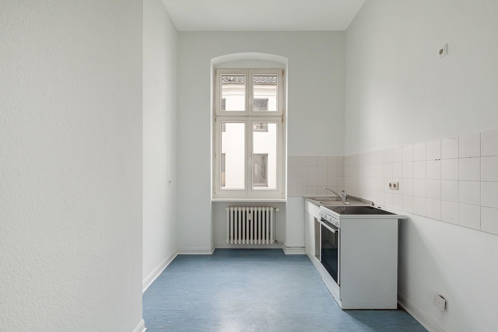 Schmale, helle Küche mit Fenster, Herd, Spüle und weißen Fliesen an der Wand.
