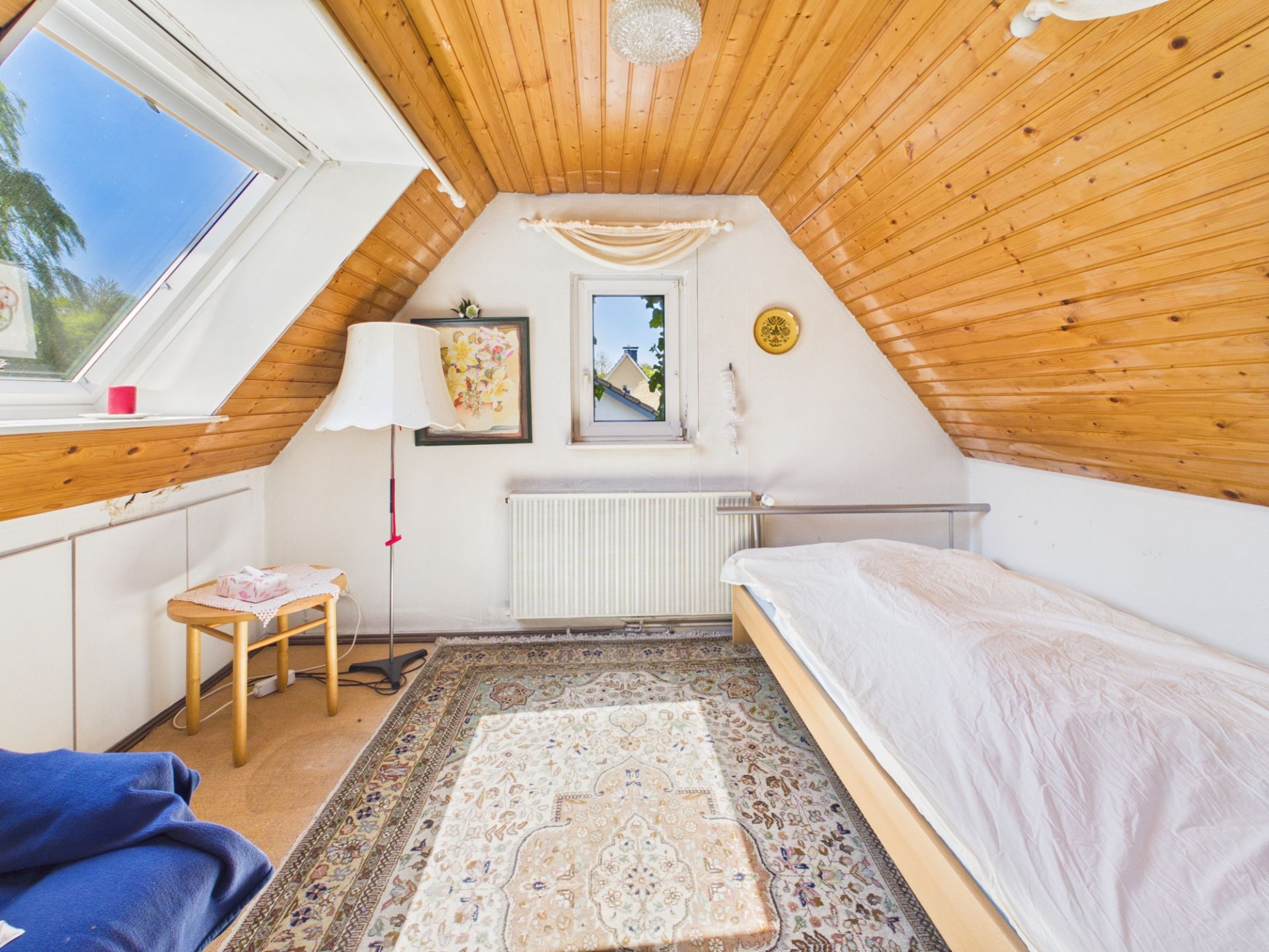 Helles Schlafzimmer mit Holzdecke, Einzelbett, Teppich und Fenster in der Dachschräge.