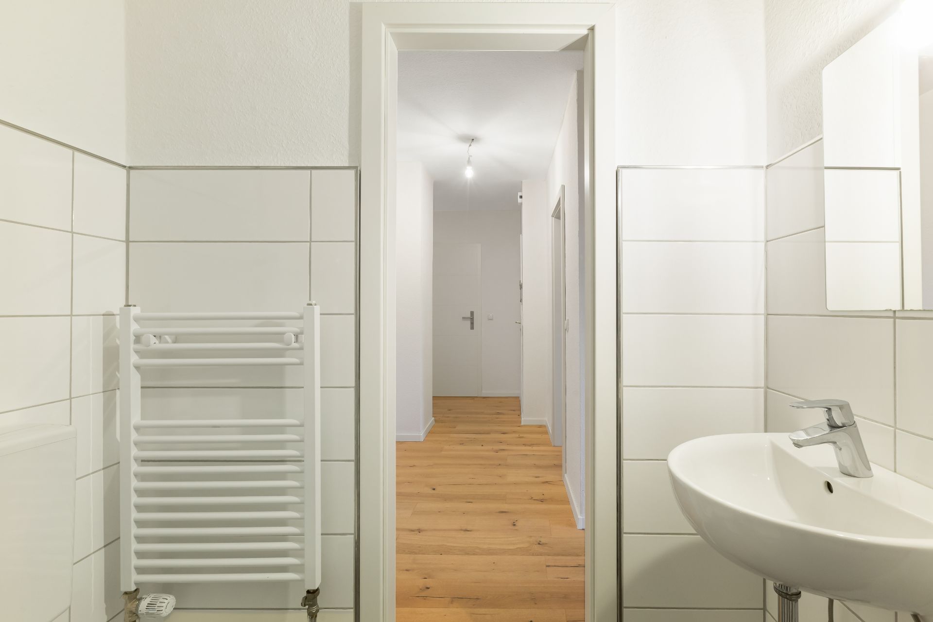 Badezimmer mit Waschbecken, Handtuchheizkörper und Blick in einen Flur mit Holzboden.