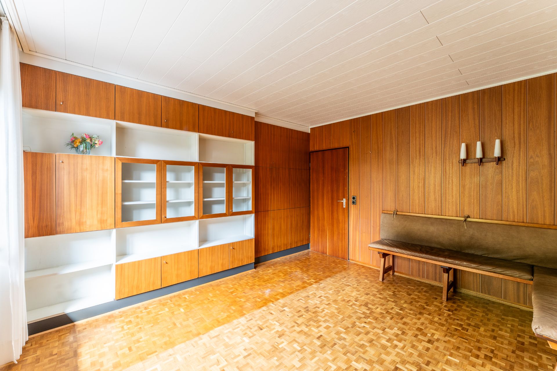 Wohnzimmer mit Holzvertäfelung, Regalwand, Bank und Blumen in einer Vase.