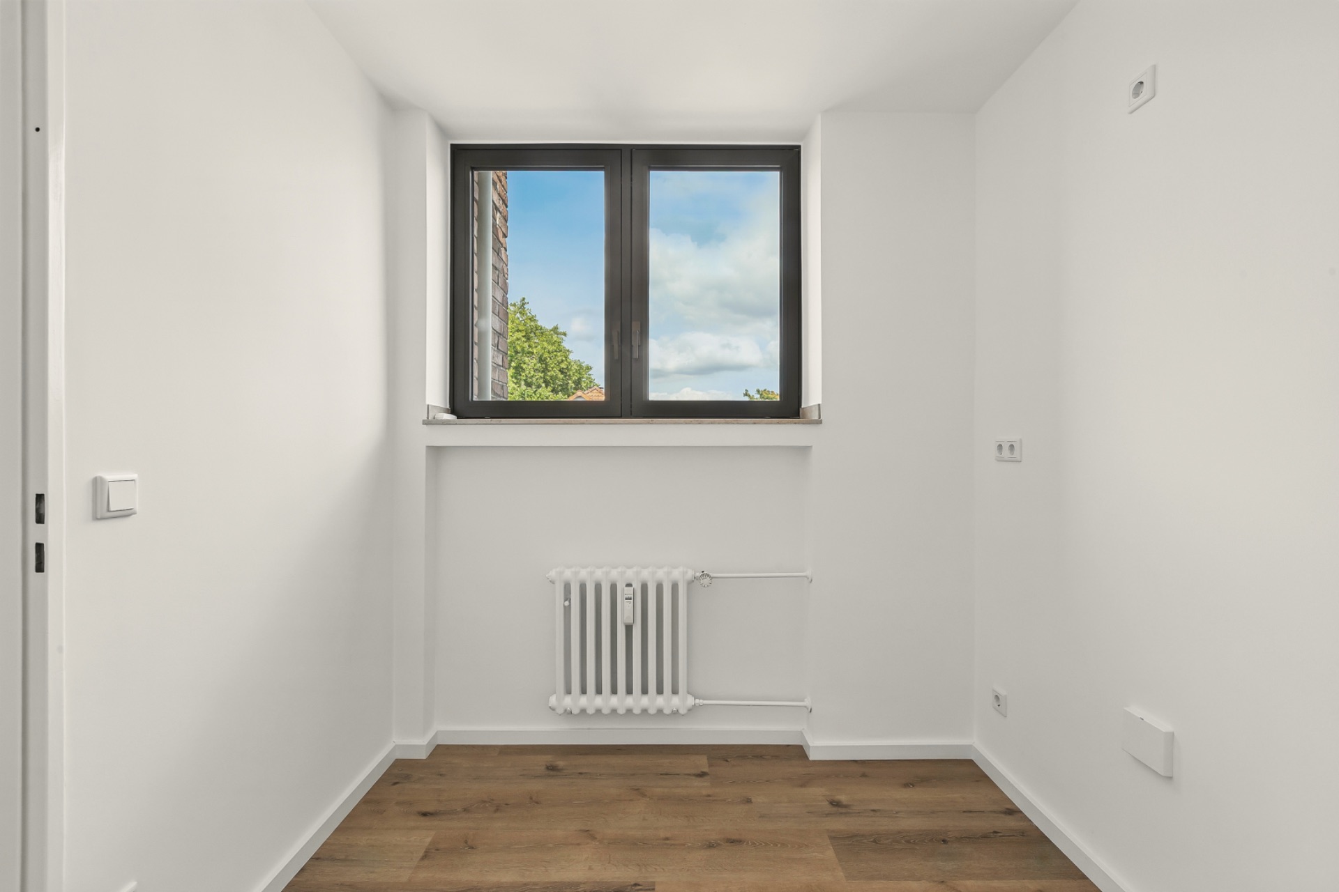 Kleiner, leerer Raum mit Holzfußboden, weißem Heizkörper und Fenster mit Blick ins Freie.
