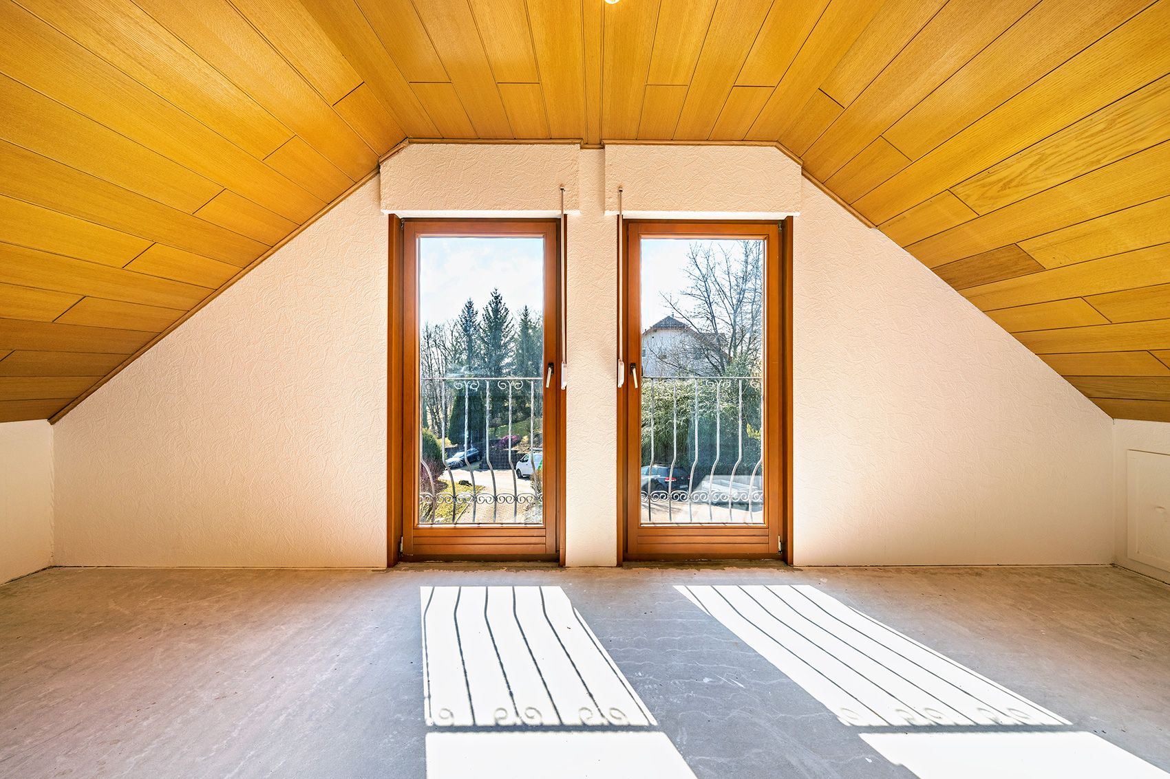 Zimmer mit schräger Holzdecke und zwei großen Fenstern mit Blick nach draußen.