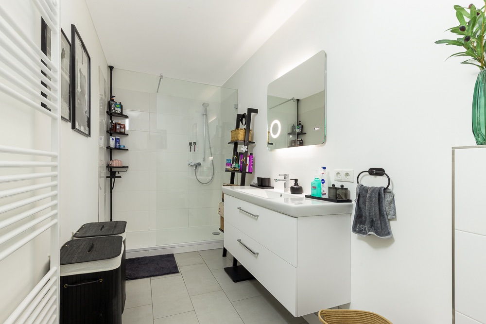 Modernes Badezimmer mit Dusche, Waschbecken, großem Spiegel und weißen Wänden.