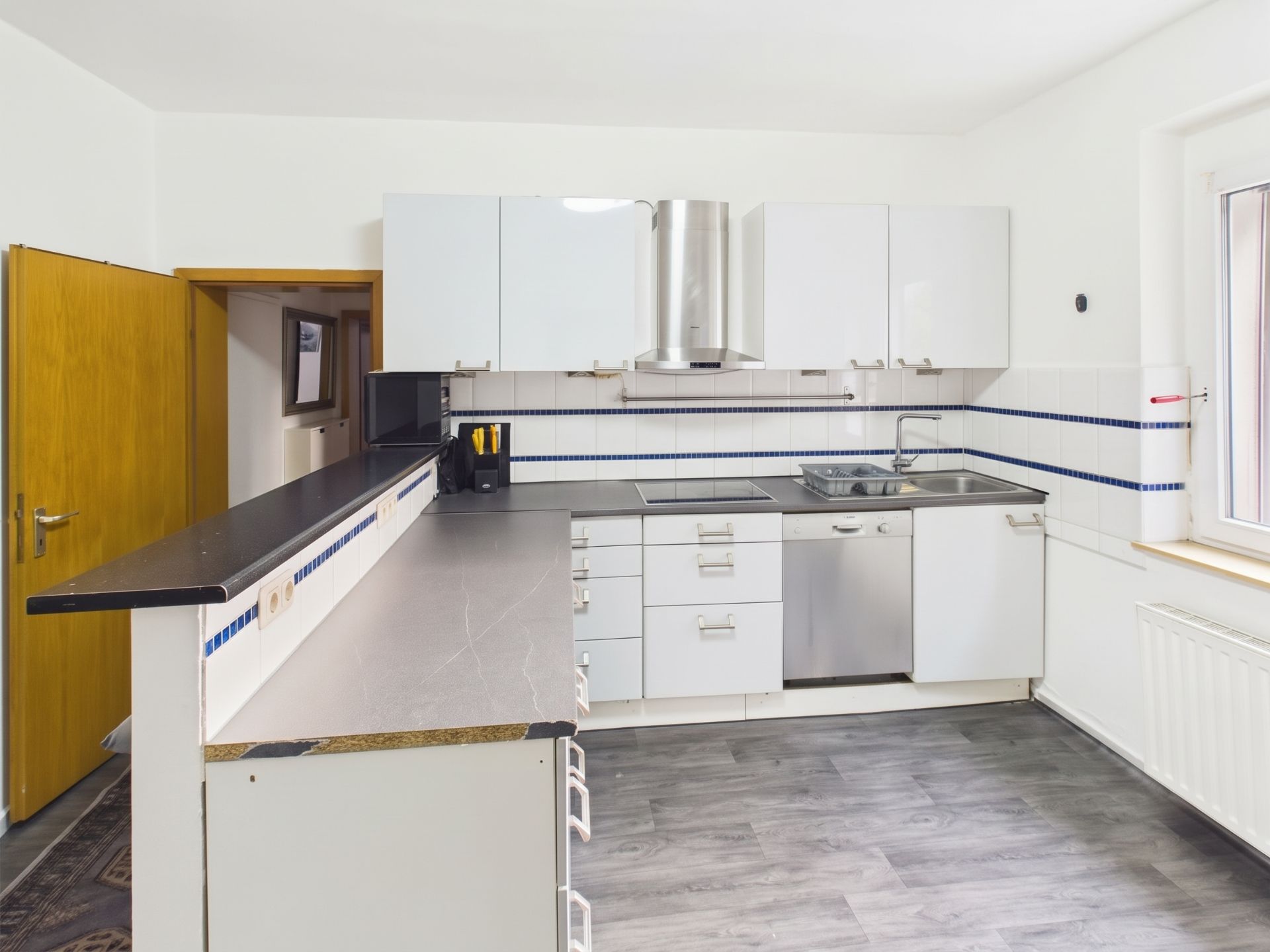 Moderne Küche mit weißen Schränken, Elektrogeräten und grauem Boden, Fenster rechts.