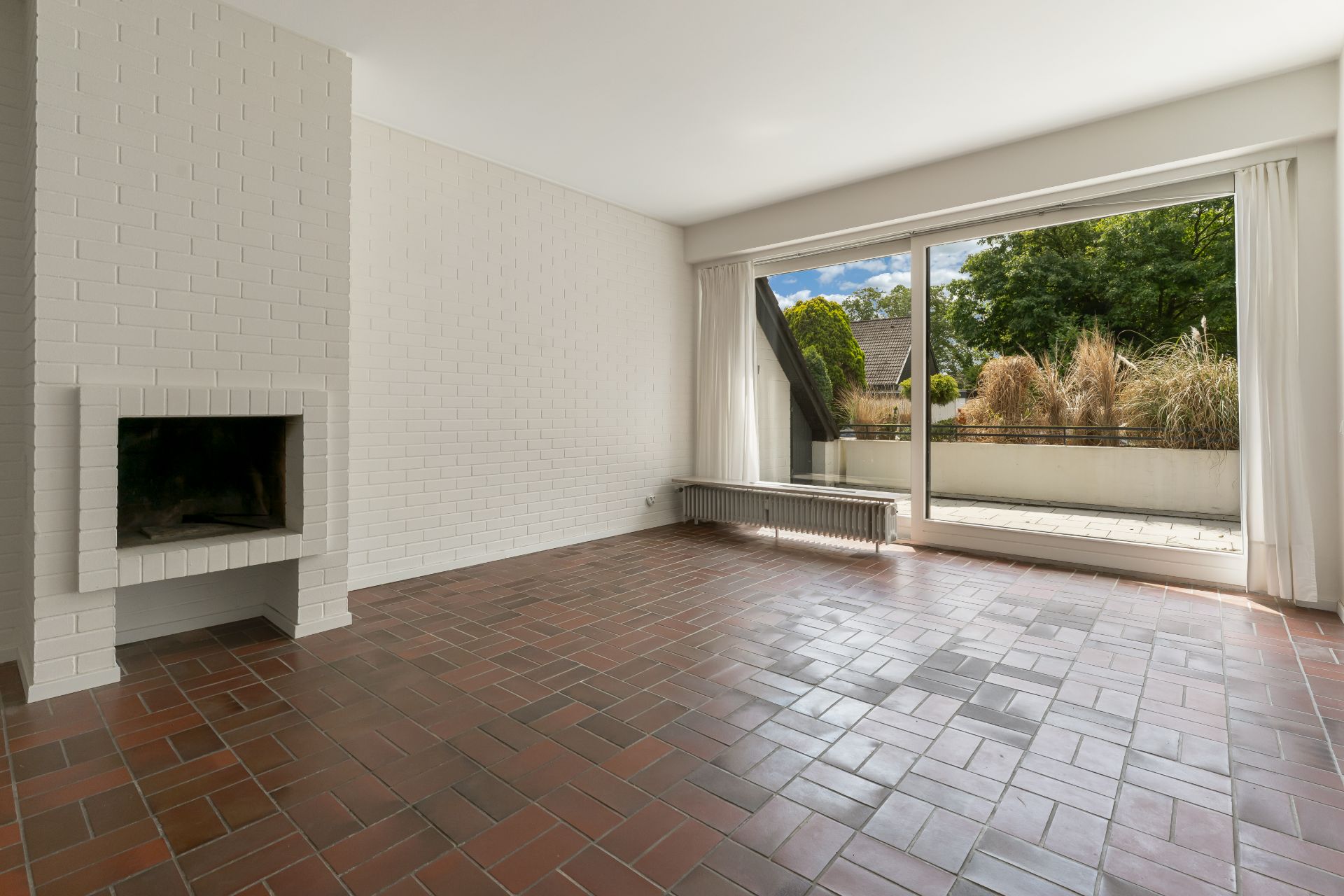 Leeres Wohnzimmer mit Kamin, braunem Fliesenboden und großer Fensterfront mit Blick auf Terrasse.