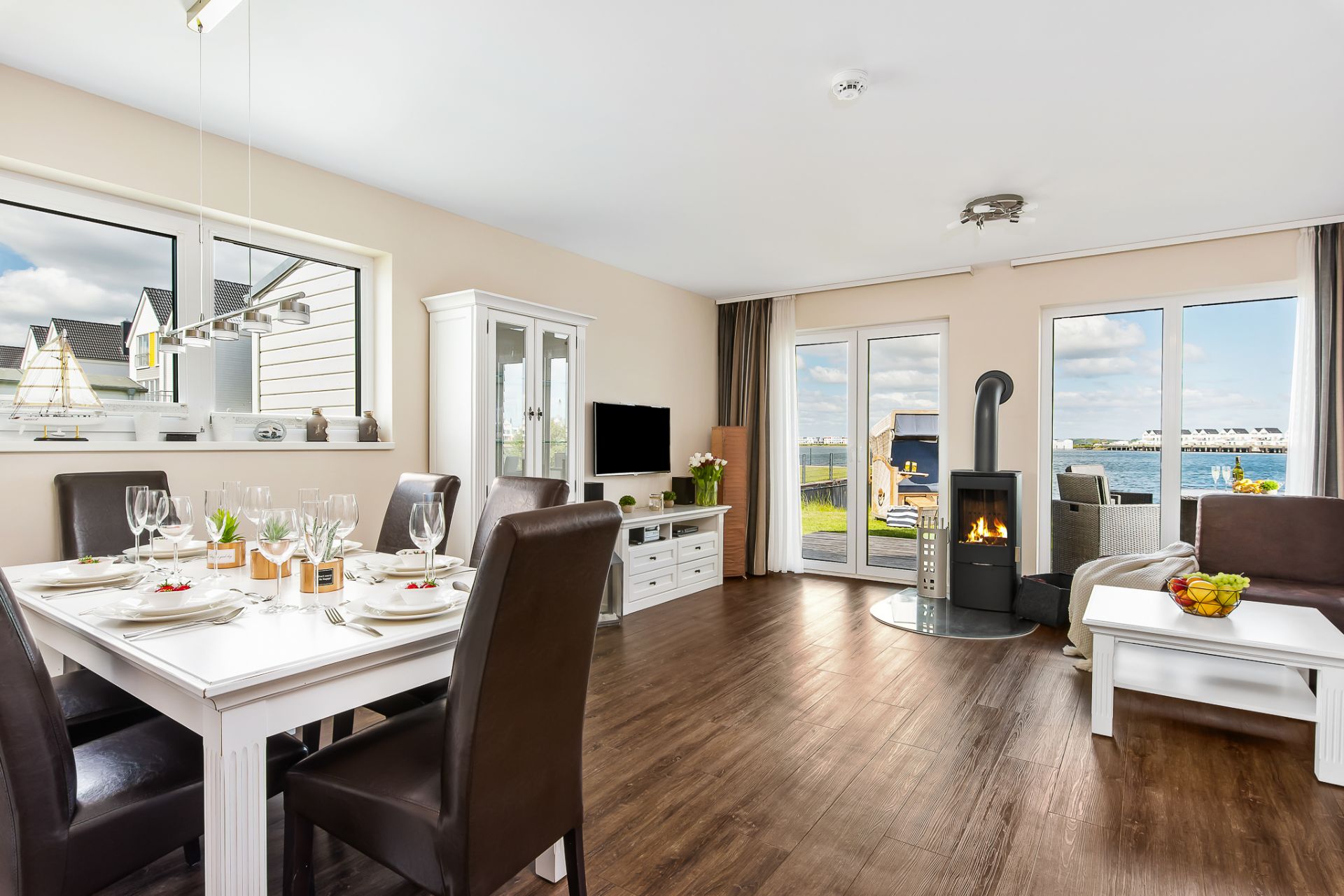 Helles Wohnzimmer mit Esstisch, Kamin, Sofa, Fernseher und Blick auf einen See durch große Fenster.